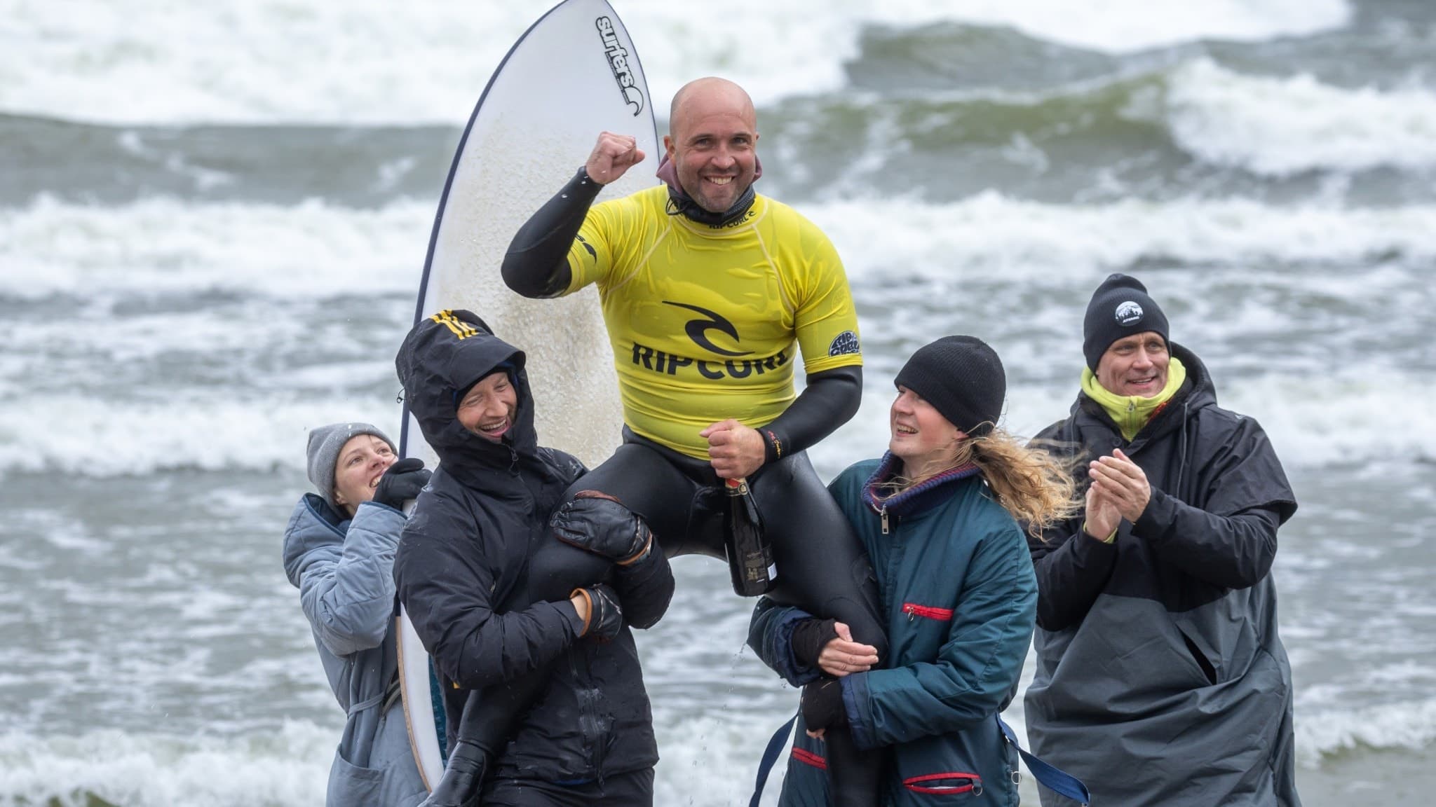 Nick Holmgren lyfts upp av ett team vid en strand efter en framgångsrik prestation i en surftävling. Han bär en gul tävlingströja och håller en näve i luften medan vågor slår mot stranden.