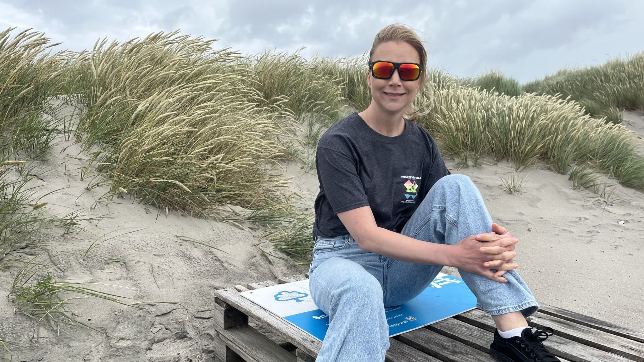 Person sitter på en träbänk vid en sandig strandmiljö med grönskande buskar i bakgrunden. Personen bär jeans och en svart t-shirt. Molnig himmel syns ovanför.