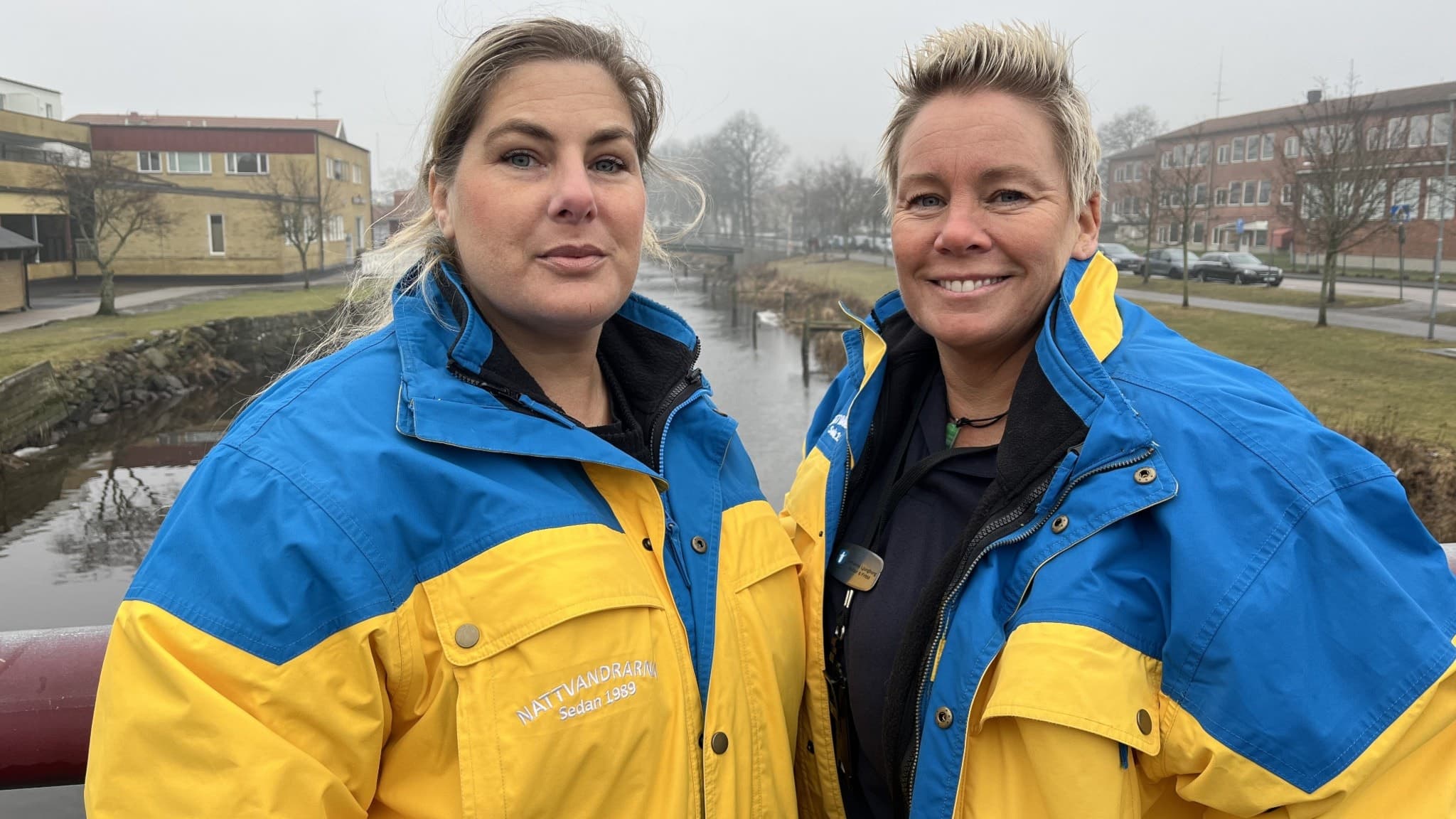 Två kvinnor i blågula jackor står på en bro över ett vattendrag med hus i bakgrunden.