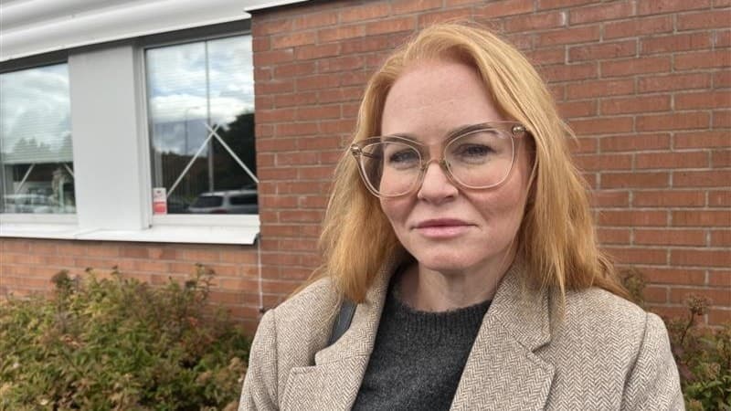 Josefine Palangi Sjöstrand står framför en tegelvägg med en fönsterrad i bakgrunden, klädd i en grå kavaj och glasögon.