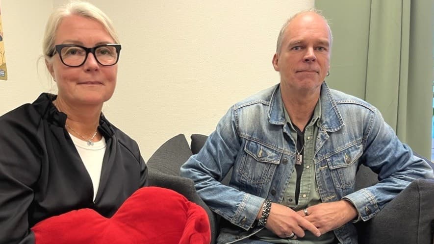 Två personer sitter i en soffa på Barnahus Halland, en håller i en röd hjärtformad kudde. Väggarna har barnteckningar och en affisch. Miljön är varm och inbjudande.