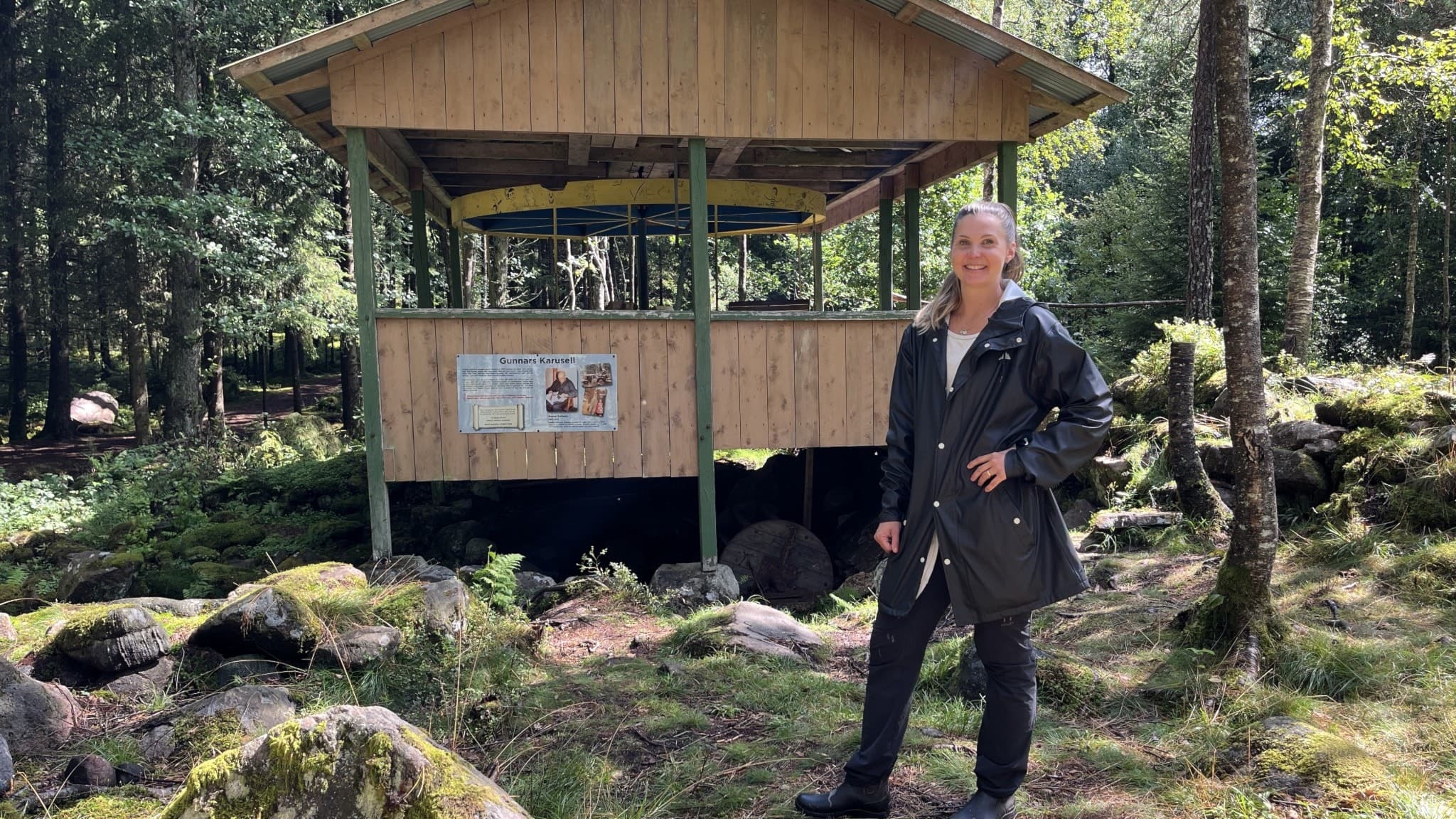 Kvinna i svart regnjacka som står framför en karusell med tak mitt ute i skogen. Unik karusell mitt i skogen får ny konstnärlig touch