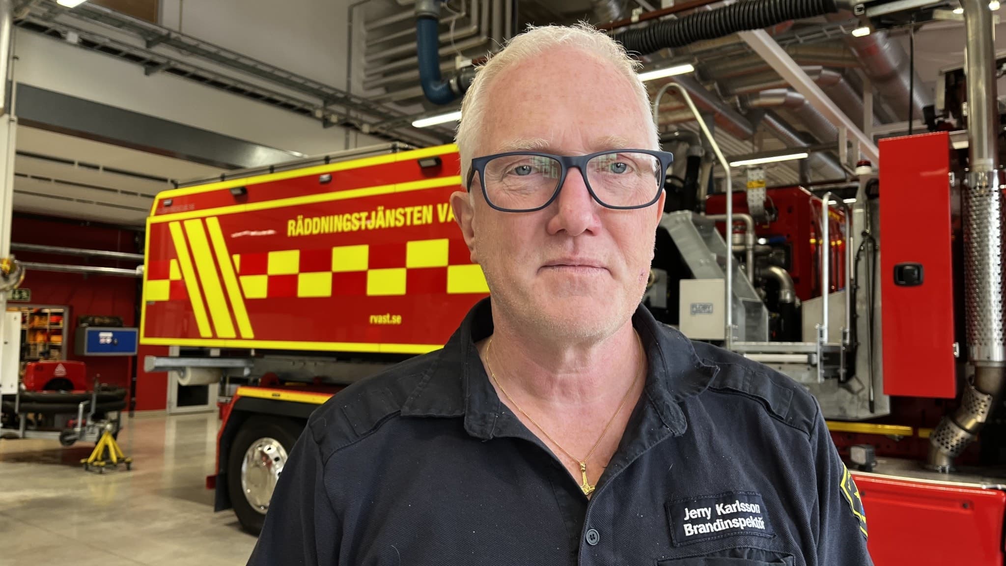 En person står framför en brandbil med texten 'Räddningstjänsten' tydligt synlig. Bilden är tagen inomhus i en brandstation med modern utrustning och industriell miljö.