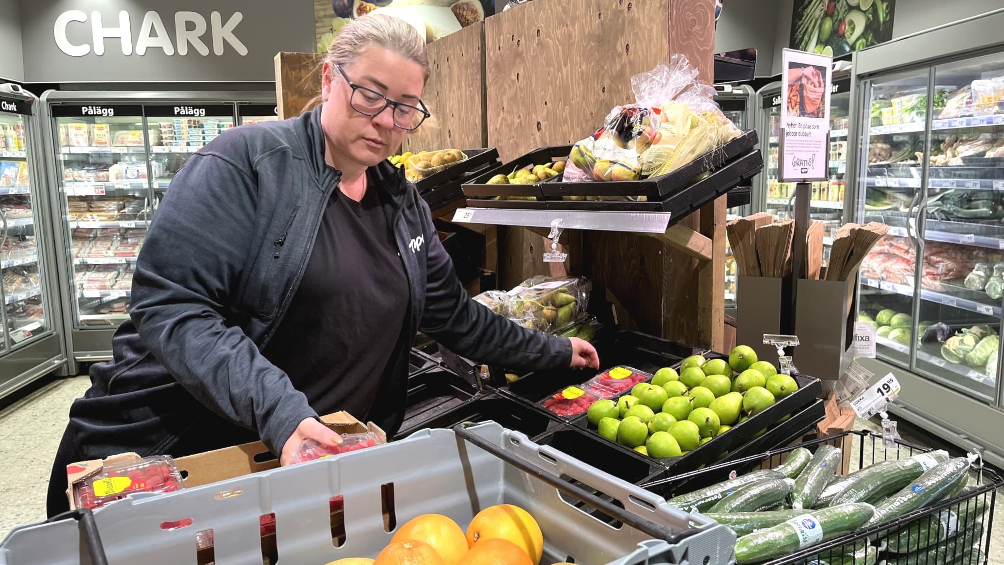 En butiksanställd i mörka arbetskläder fyller på färska gröna äpplen vid en fruktavdelning i en livsmedelsbutik. I bakgrunden syns kylskåp med köttprodukter och andra matvaror.