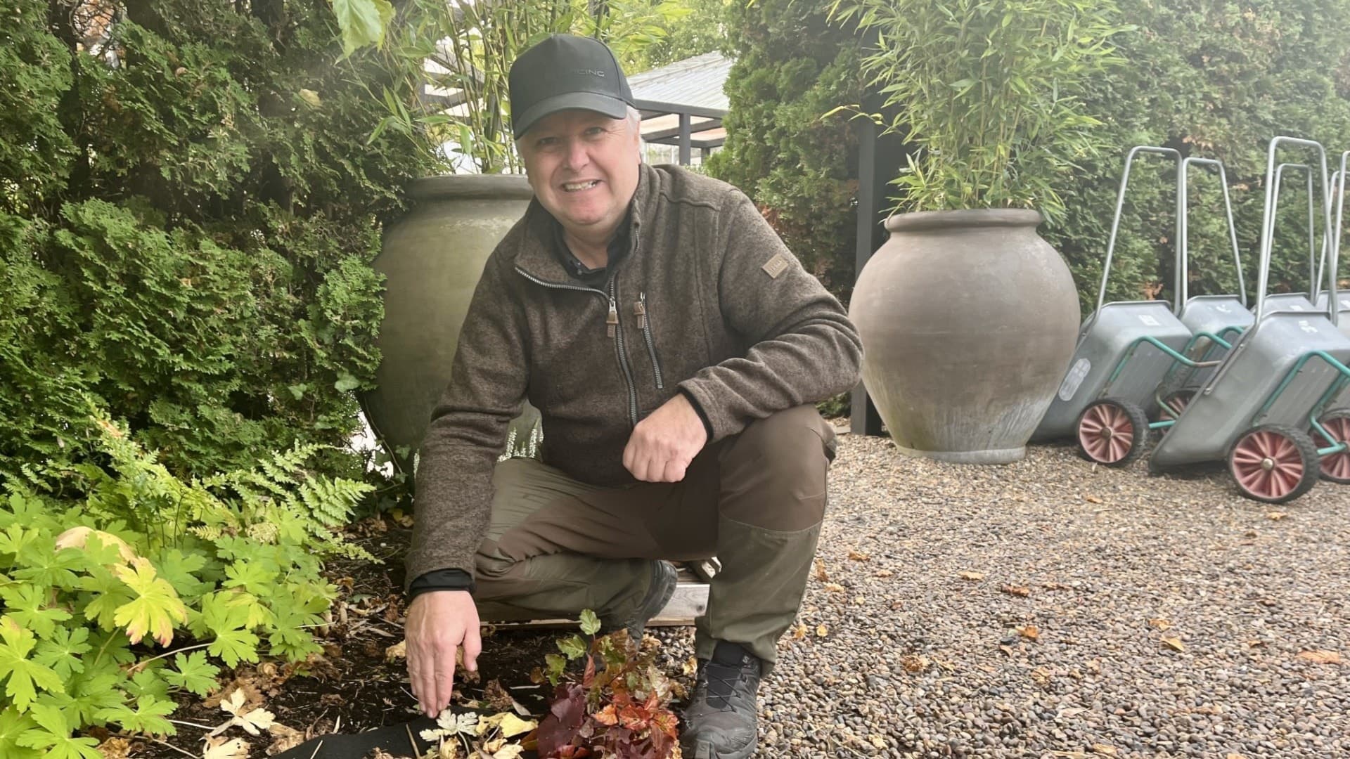 Trädgårdsmästaren Lars-Johan Svanström sitter på huk i en grönskande trädgård, omgiven av växter, stora krukor och skottkärror på en grusgång.