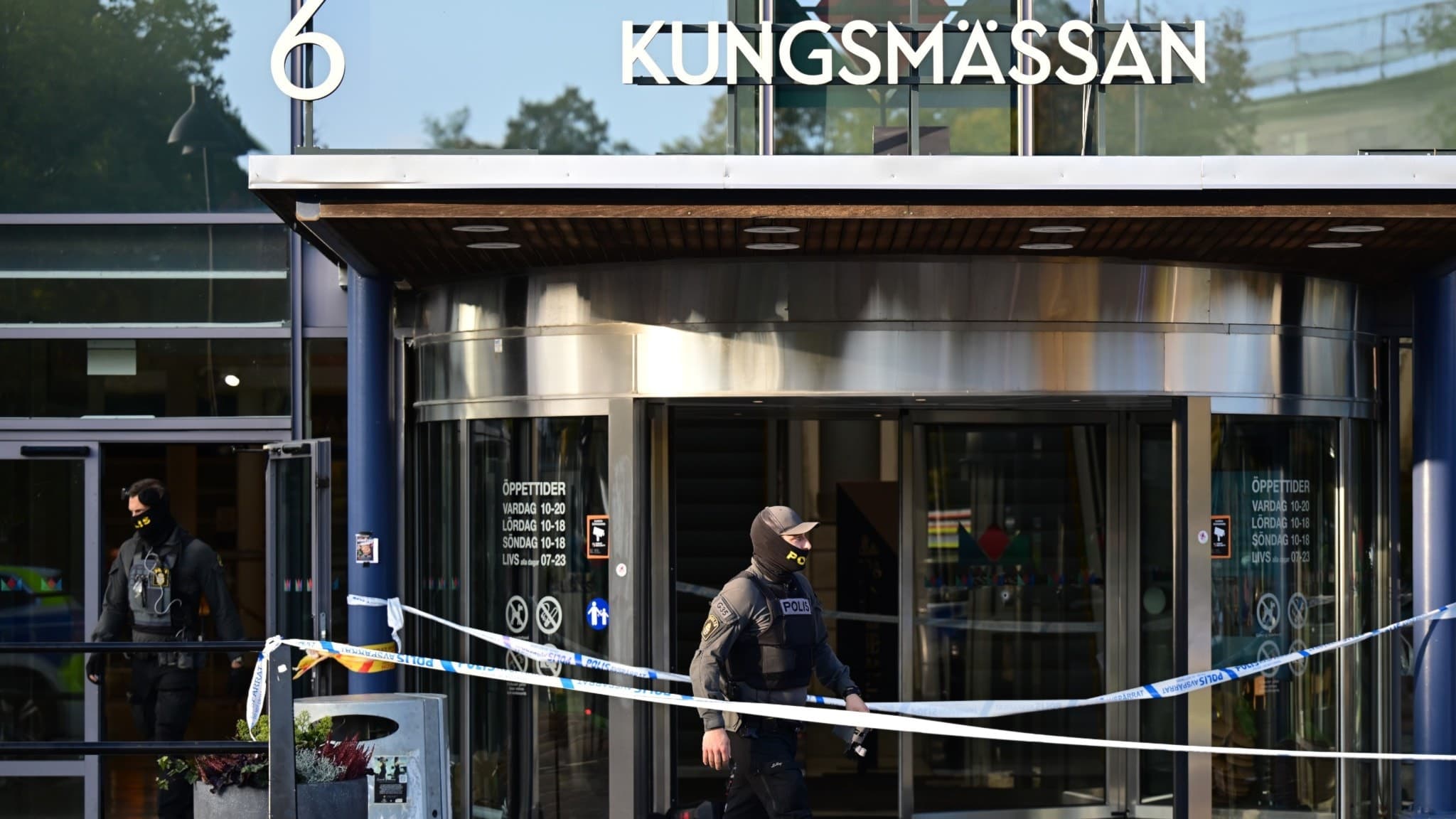 Entrén till Kungsmässan avspärrad med polisband efter en skottlossning. En maskerad polis rör sig vid platsen. Glasdörrar och skylt med texten 'Kungsmässan' syns i bakgrunden.