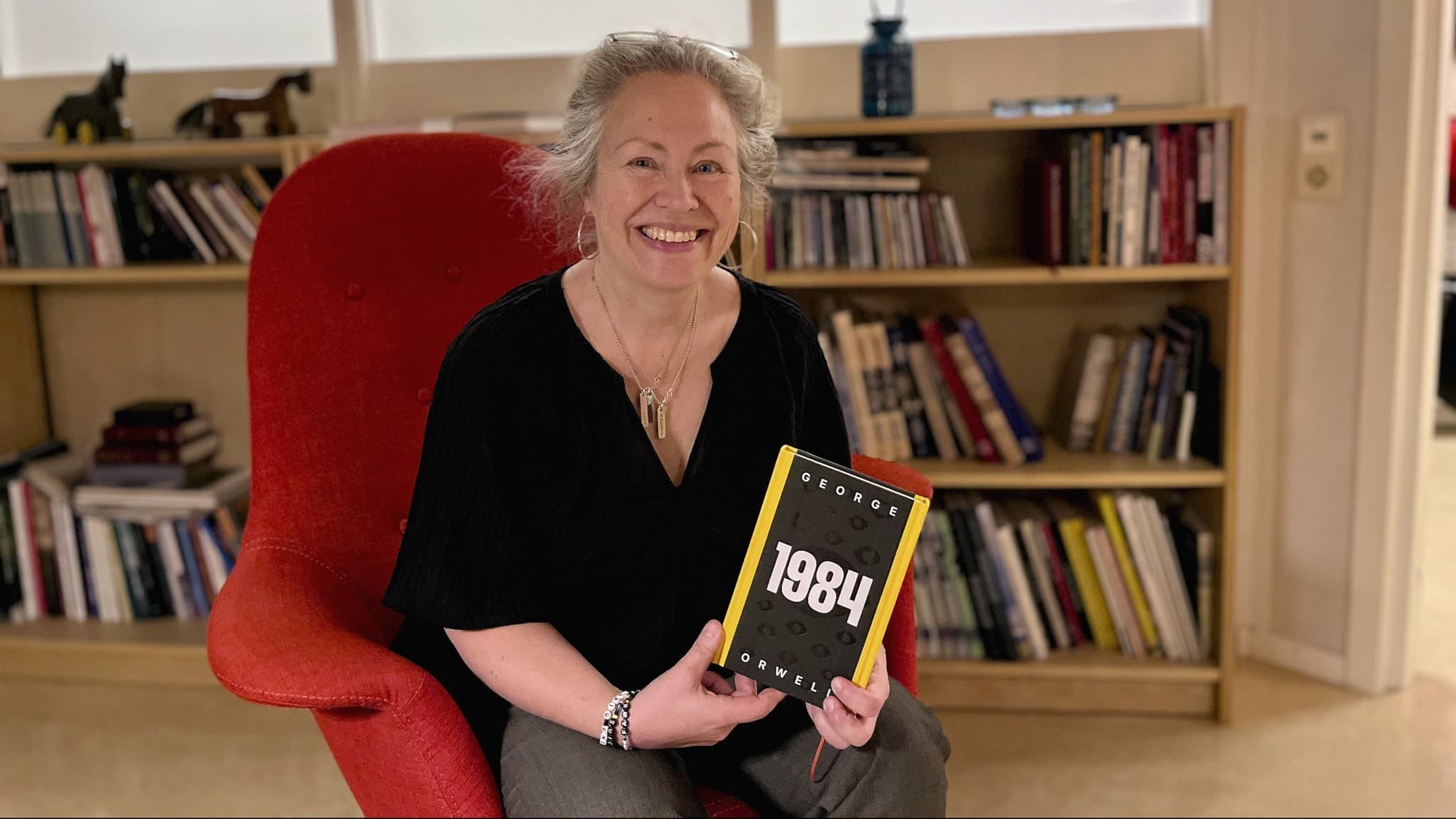 Ulrika Ahlberg sitter i en röd stol och håller upp en bok med titeln 'Banned books' framför en bokhylla i ett bibliotek. Bilden illustrerar temat förbjudna böcker.