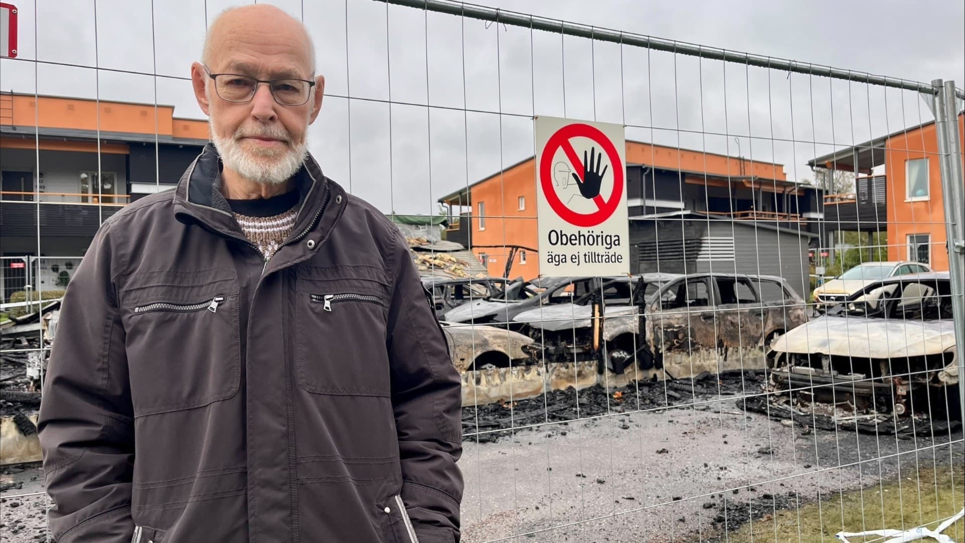En man står framför ett stängsel med flera utbrända bilar i bakgrunden, inklusive Anders Johanssons bil som totalförstördes i en misstänkt mordbrand i Hallsberg. En skylt förbjuder obehöriga.