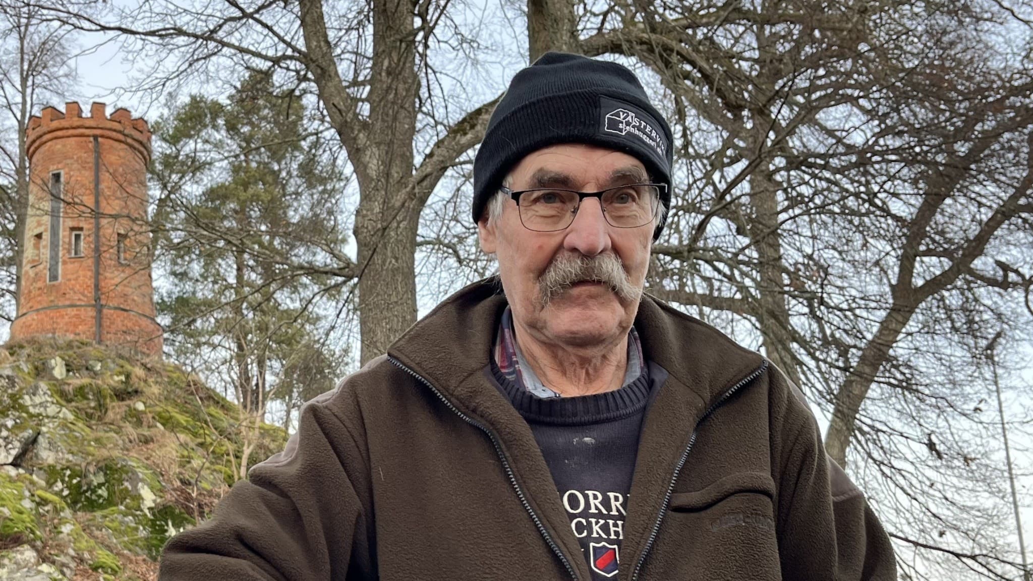 Lars Rasch, ordförande i PRO  i Åtvidaberg, står utomhus framför ett historiskt torn omgivet av träd. Han är klädd i mörka vinterkläder och bär en mössa.