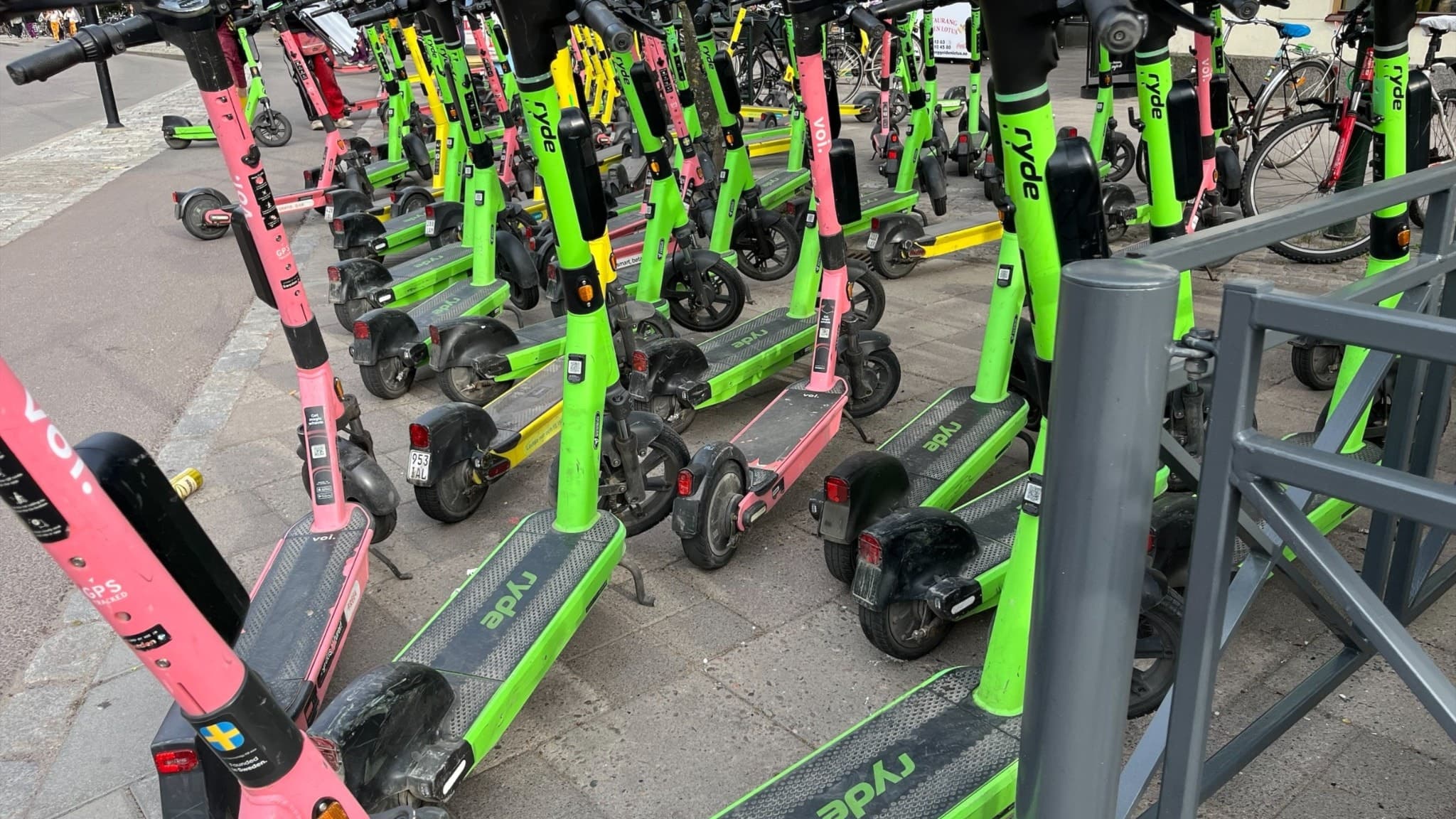 Flera elsparkcyklar i olika färger, bland annat rosa och gröna, står parkerade i en tät rad på en trottoar i en stadsmiljö.