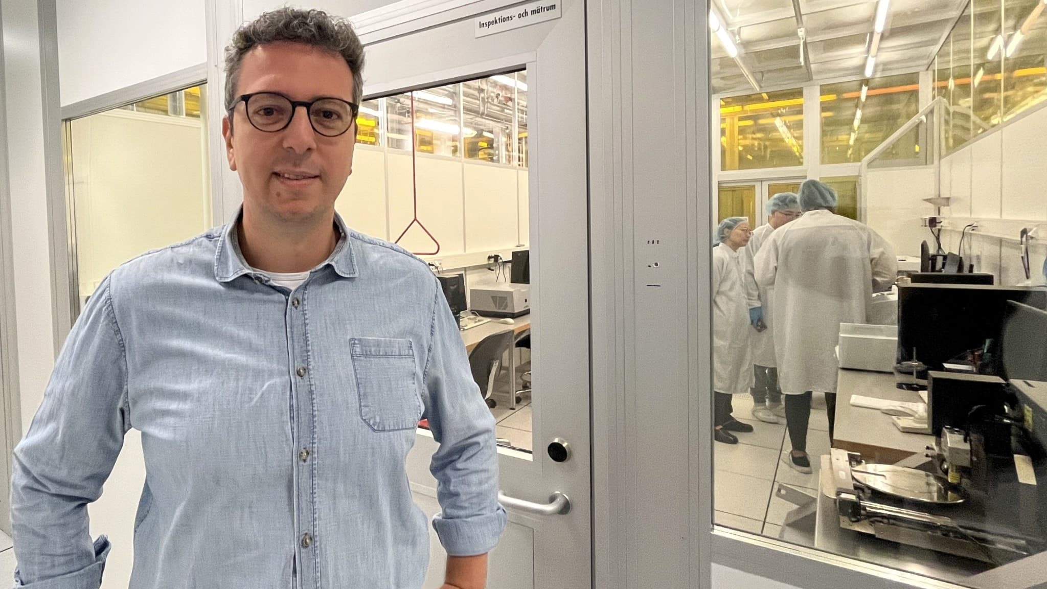 Simone Fabiano, professor vid LiU, står i en modern laboratoriemiljö med glasväggar och avancerad teknisk utrustning synlig i bakgrunden. Atmosfären signalerar forskning och innovation. Det de forskar på är om det går att ta fram artificiella nervceller för att göra implantat som ska kunna läsa av nivåer i kroppen och rummet och sedan larma och agera.