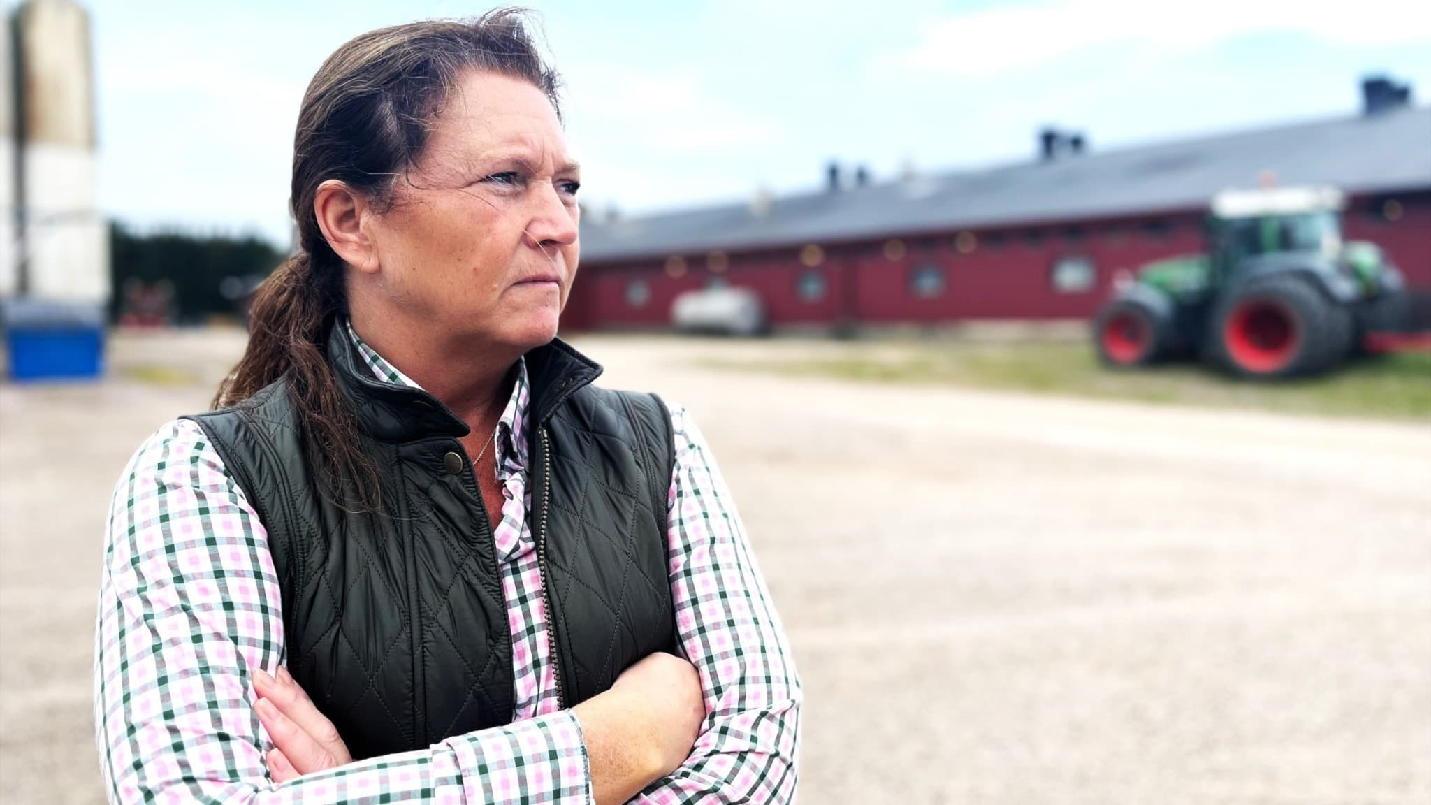 Jeanette Blackert, ordförande i LRF Östergötland, står på en gårdsplan med en röd lada och en grön traktor i bakgrunden, symboliserande lantbruk och lokal jordbruksverksamhet.