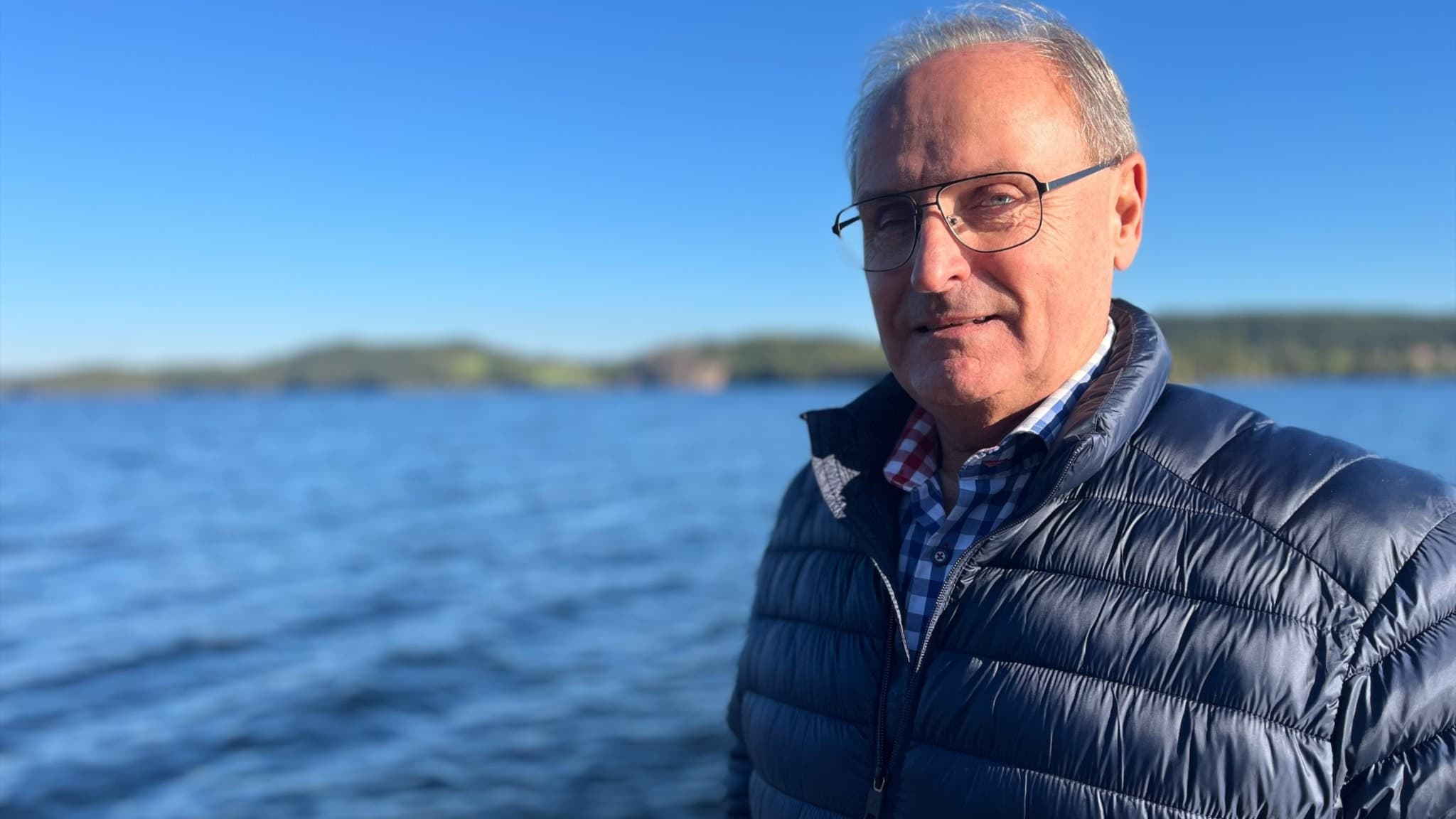 Tomas Johansson, ordförande för föreningen Ydre näringsliv, står vid en sjö med glittrande blått vatten och skog i bakgrunden under en solig himmel.