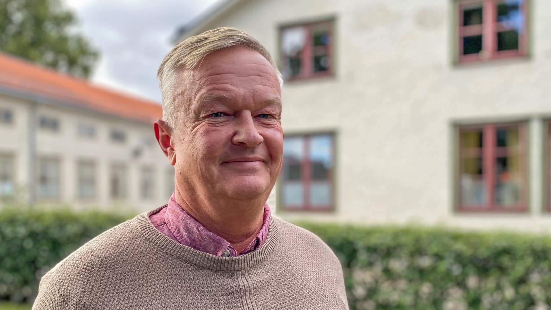 Man med blont kort hår som ler. Stickad tröja där en rosa skjorta sticker upp. 