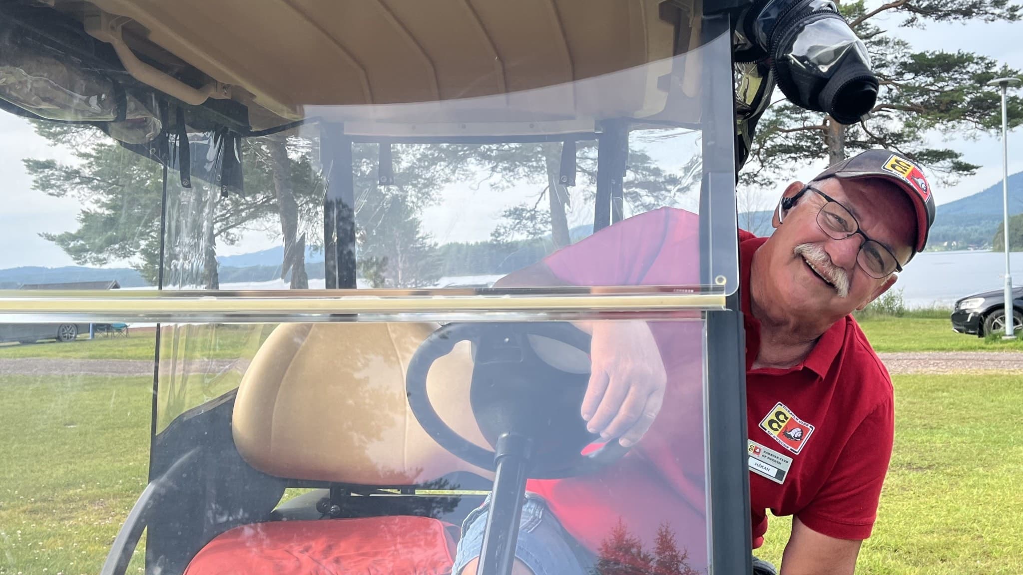 Campingvärden Håkan Zetterberg på Sollerön står vid en golfbil på en gräsplan med sjö och berg i bakgrunden. Bilden visar en campingmiljö med natur och parkerade bilar.