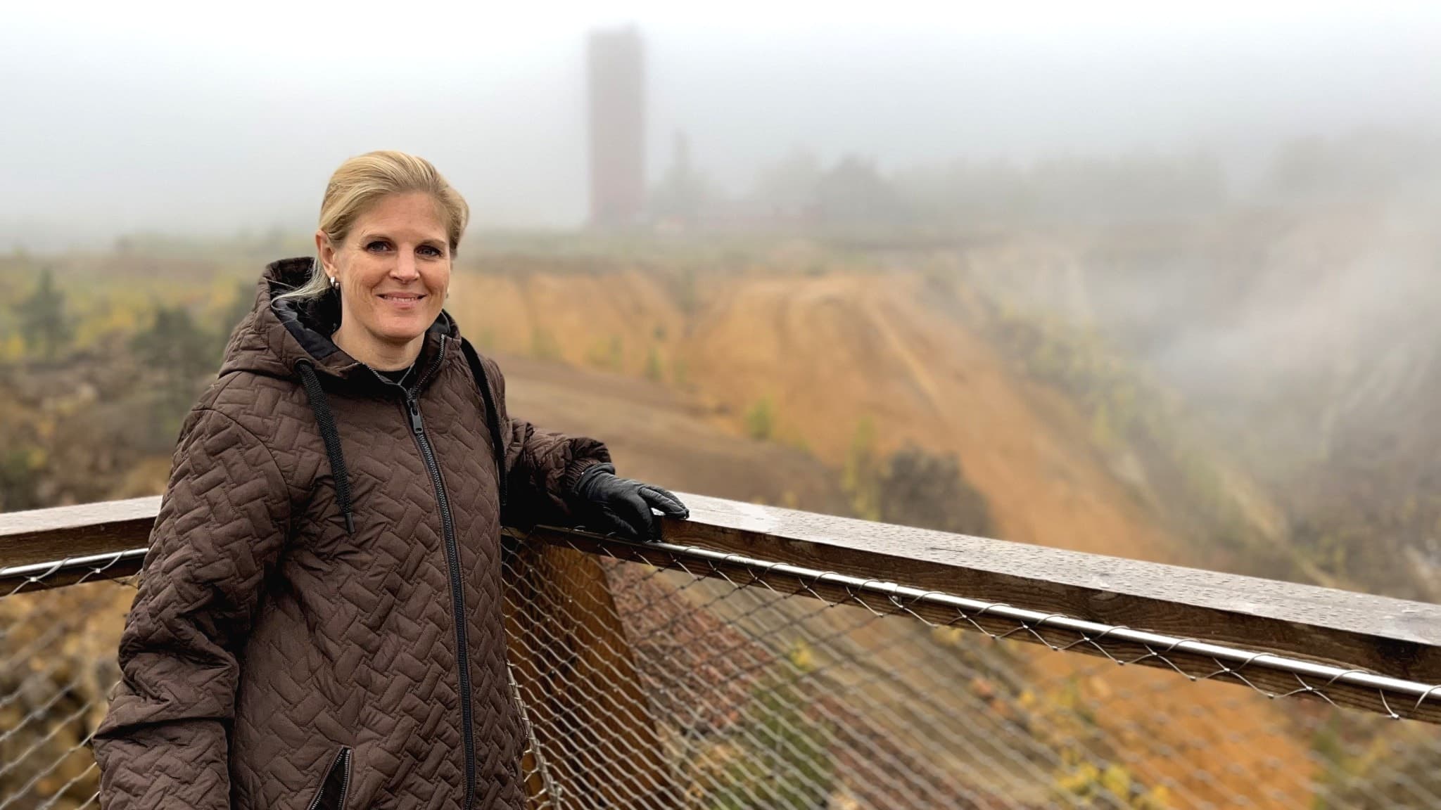 En blond kvinna står vid ett räcke med utsikt över ett dimmigt landskap med gruvområde och grönska i bakgrunden.