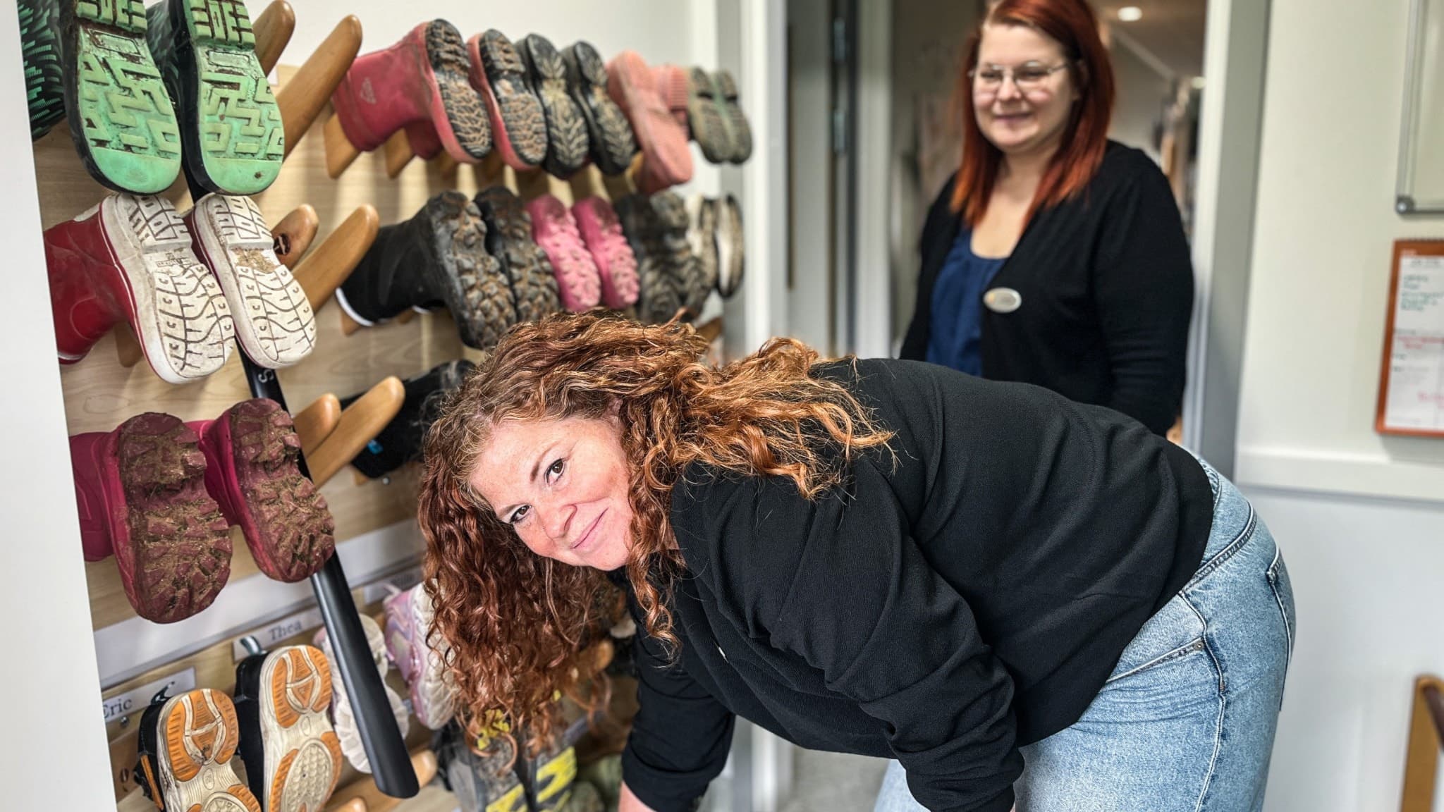 Malin Björk står vid en vägg med barnskor på en förskola i Djurås. Hon ler mot kameran och har lockigt hår, medan hyllan är fylld med färgglada skor.