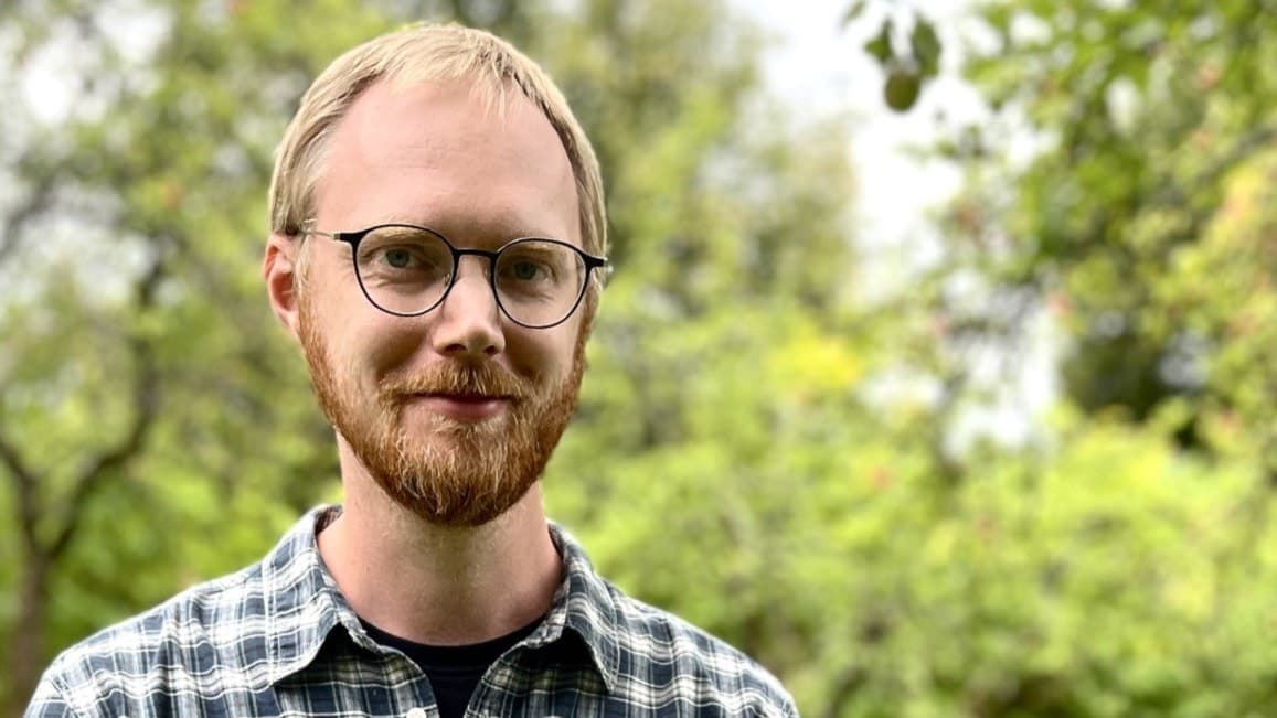 Man identifierad som David Aura står i en grönskande park, klädd i en svartvit-rutig skjorta. Träd och gräsmatta syns i bakgrunden.