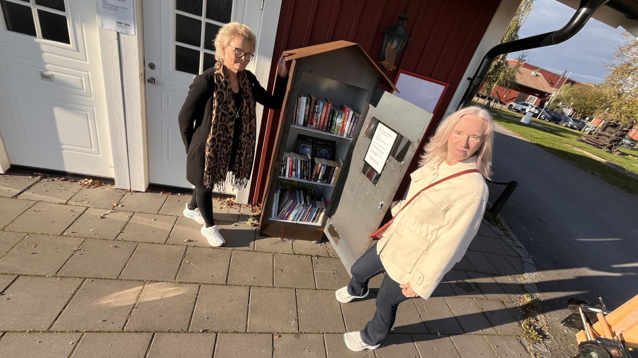 Två kvinnor står framför en röd byggnad och har öppnat ett plåtskåp som har flera hyllor med böcker.