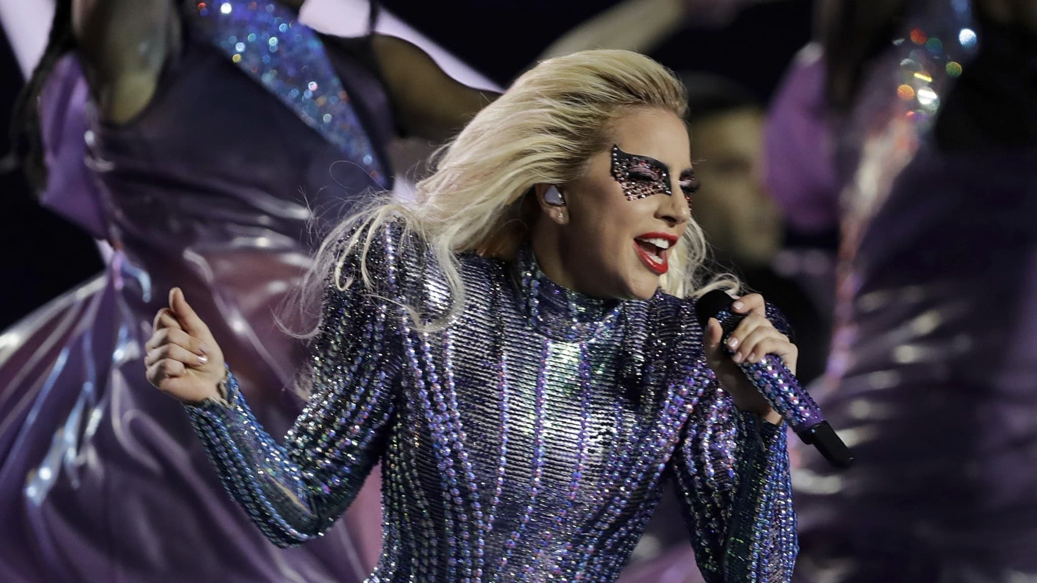 Bilden visar ett foto i närbild på Lady Gaga som står på scnen och håller i en mick. Hom har glittrig tight lila scenkläder och dekorerad med pärlor och smink runt ögonen. I ena handen håller hon en mick.
