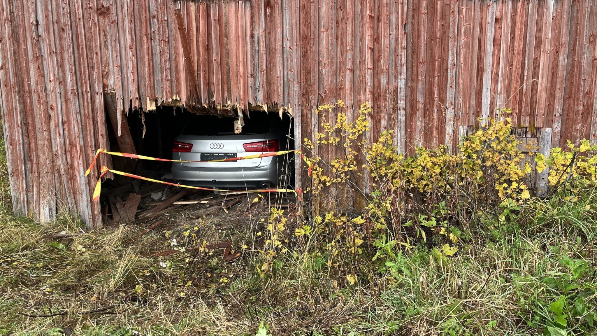 En personbil har kraschat in i en röd träbyggnad med brädvägg, vilket skapat ett stort hål. Gula markeringsband syns runt skadan och höstväxter omger byggnaden.