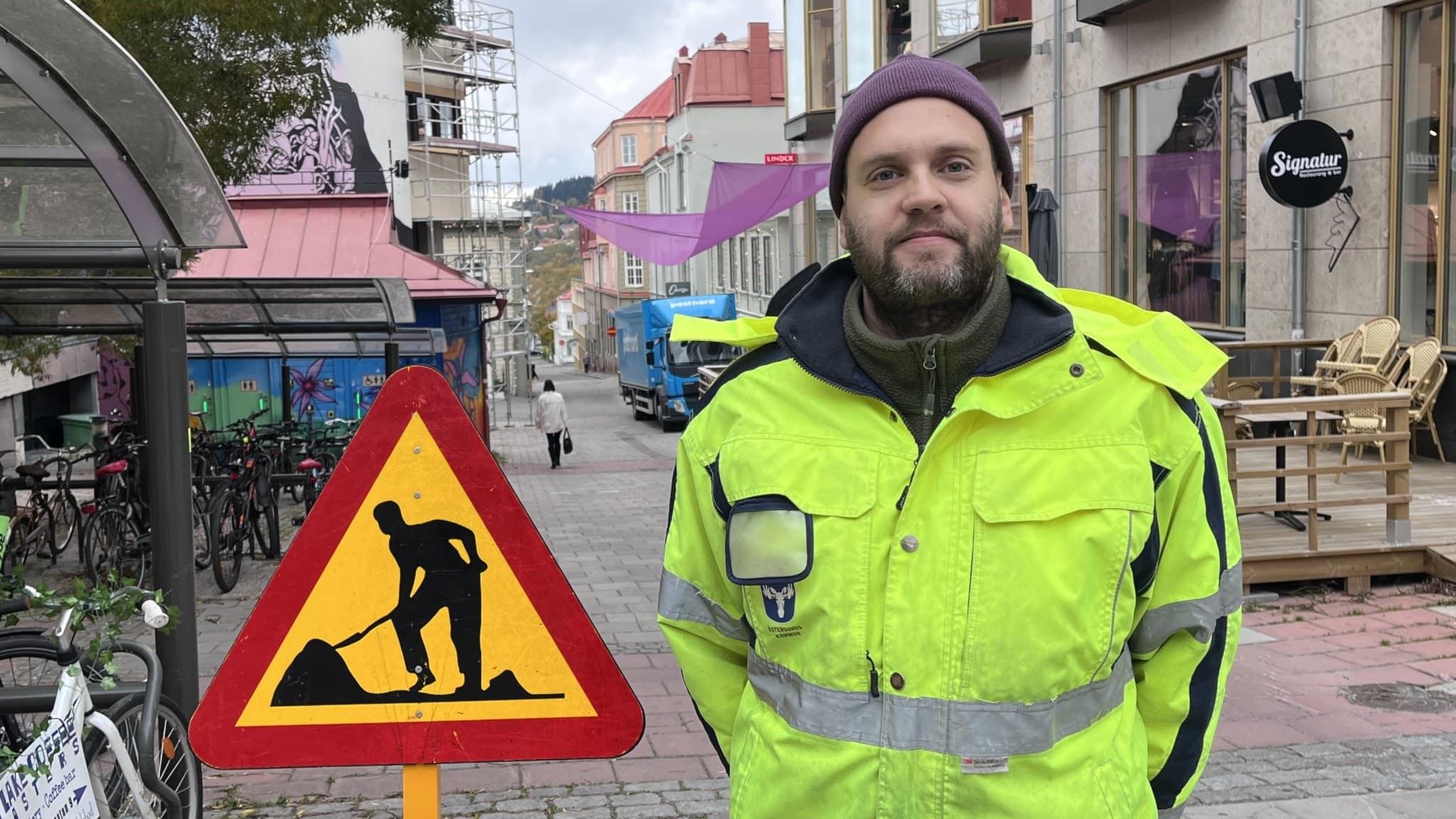 Henrik Ring, trafikhandläggare på Östersunds kommun, står utomhus i en gul reflexjacka framför ett byggarbetsområde med ett varningsskylt för vägarbete i stadsmiljö.