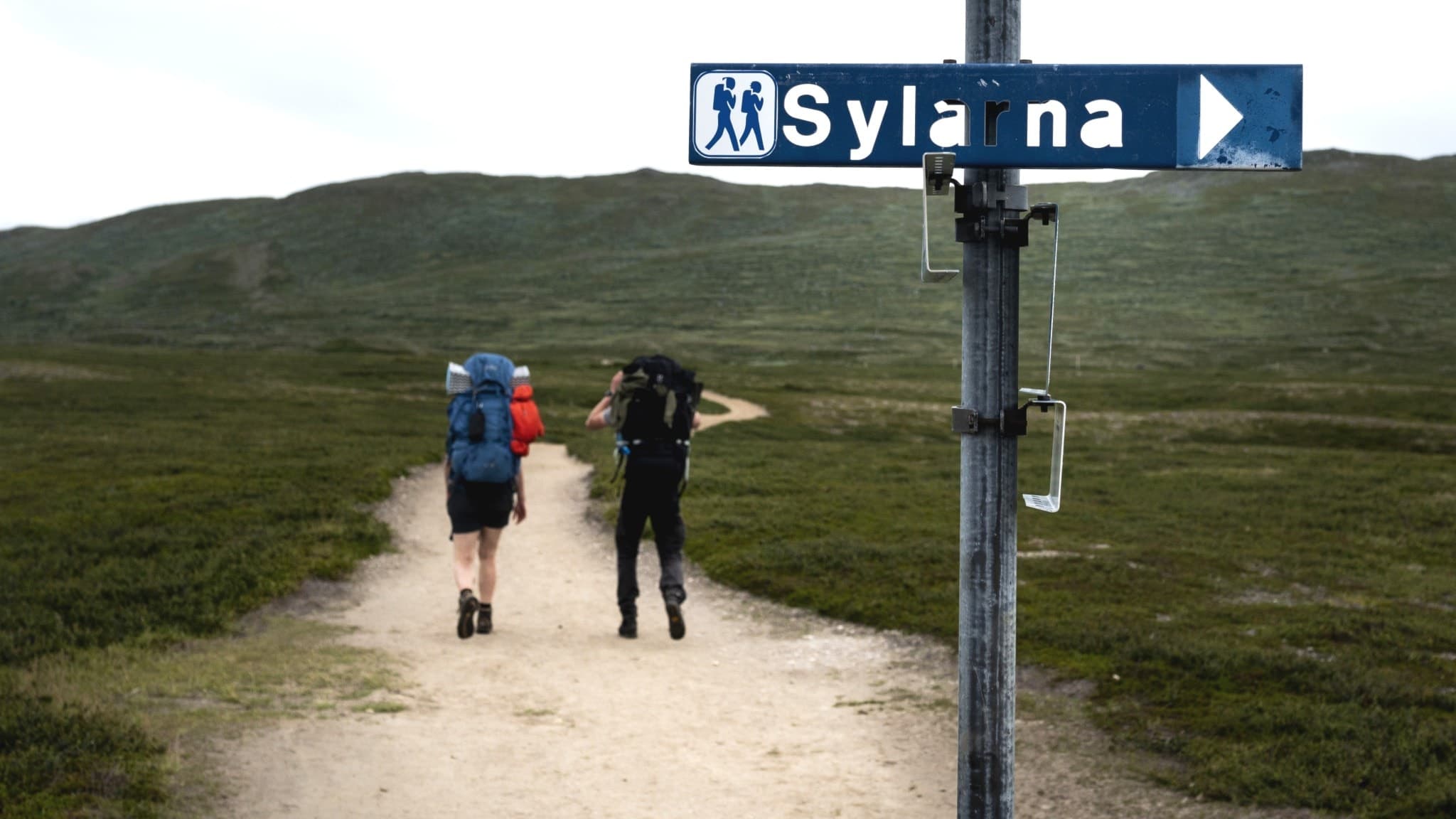 Två fjällvandrare med ryggsäckar går längs en vandringsled i fjällen med en vägvisare till Sylarna uppsatt på en stolpe