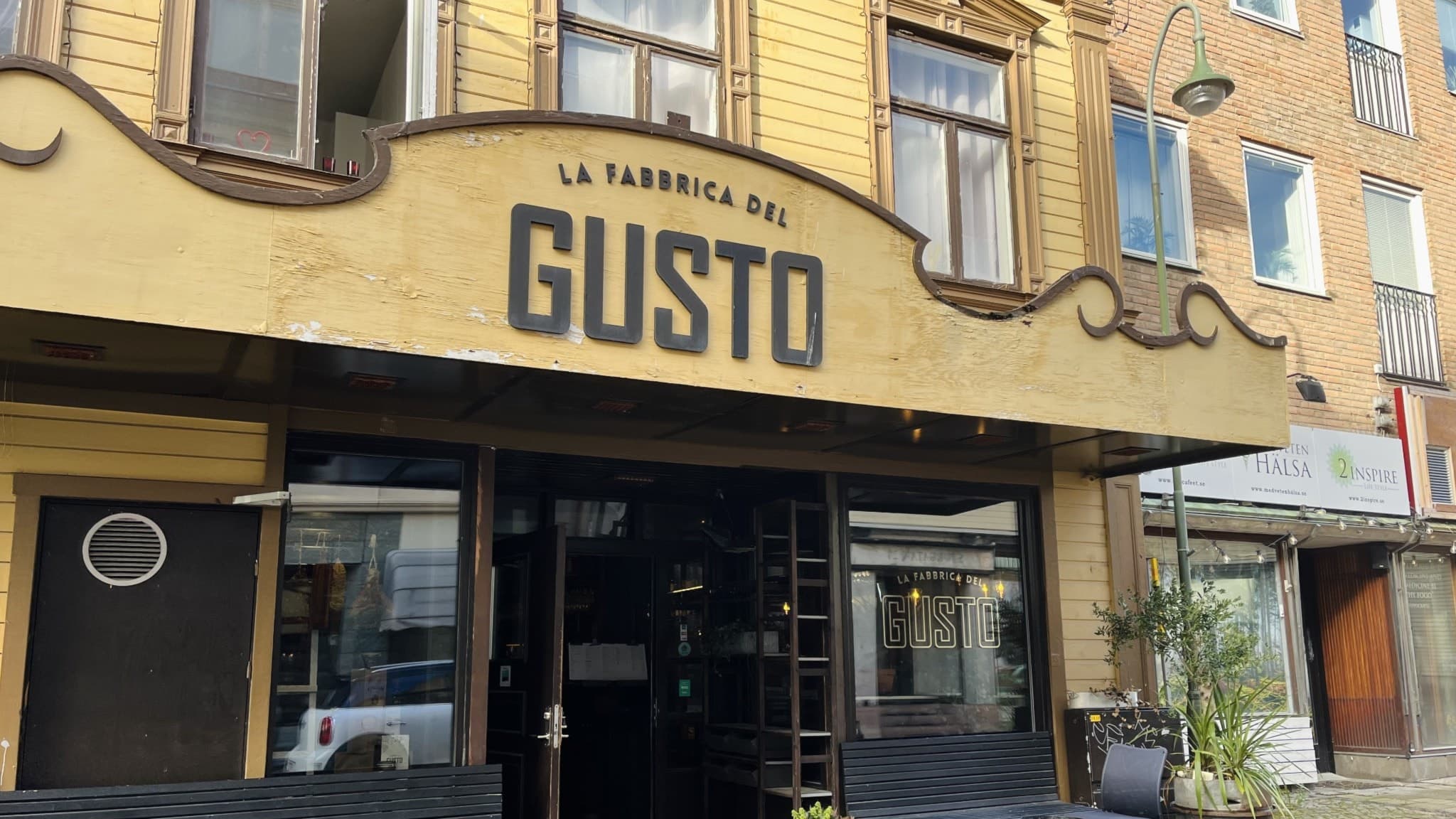 Fasaden av restaurangen La Fabrica del Gusto med gulmålade väggar och svarta utemöbler på trottoaren. Ett välvt skylt med restaurangens namn syns ovanför entrén.