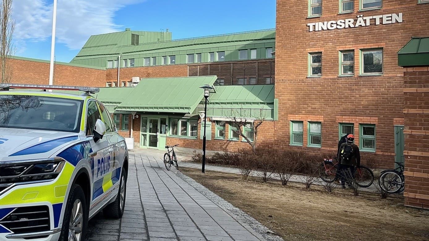 En målad polisbil i vitt, blått och gult framför en stor tegelbyggnad med grönt tak. 