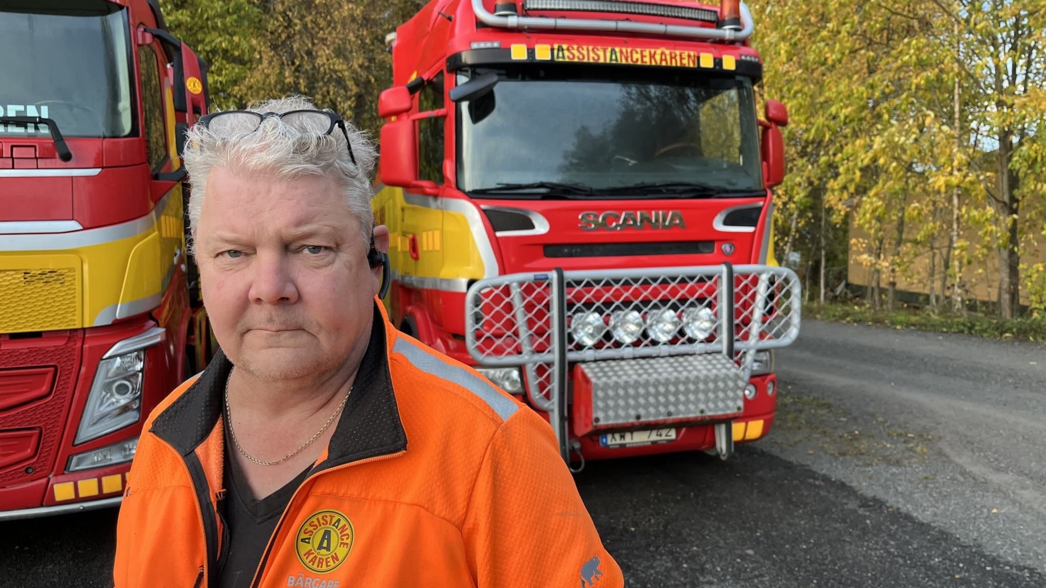 Två röda lastbilar med gula detaljer står parkerade på en landsväg, omgivna av höstträd. En person i orange arbetsjacka syns framför fordonen.