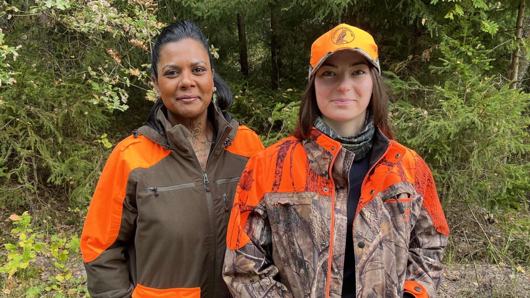 Två personer, Filippa och Nimali, iklädda jaktkläder med orange detaljer står utomhus framför grönskande skogsmiljö. Bilden framhäver jakt som naturaktivitet och deras utrustning.