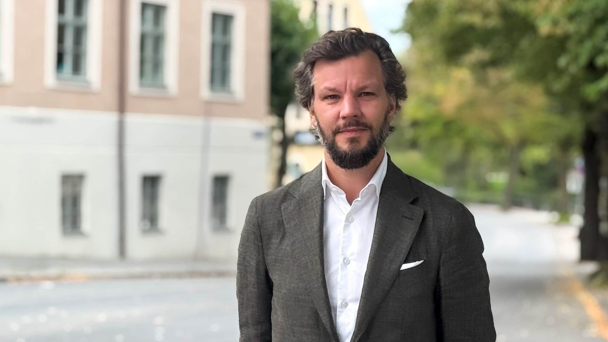 En man i grå kavaj och vit skjorta står på en tom gata med träd och byggnader i bakgrunden. Han har händerna i fickorna och ser välklädd ut. Miljön är stadsmässig och lugn.