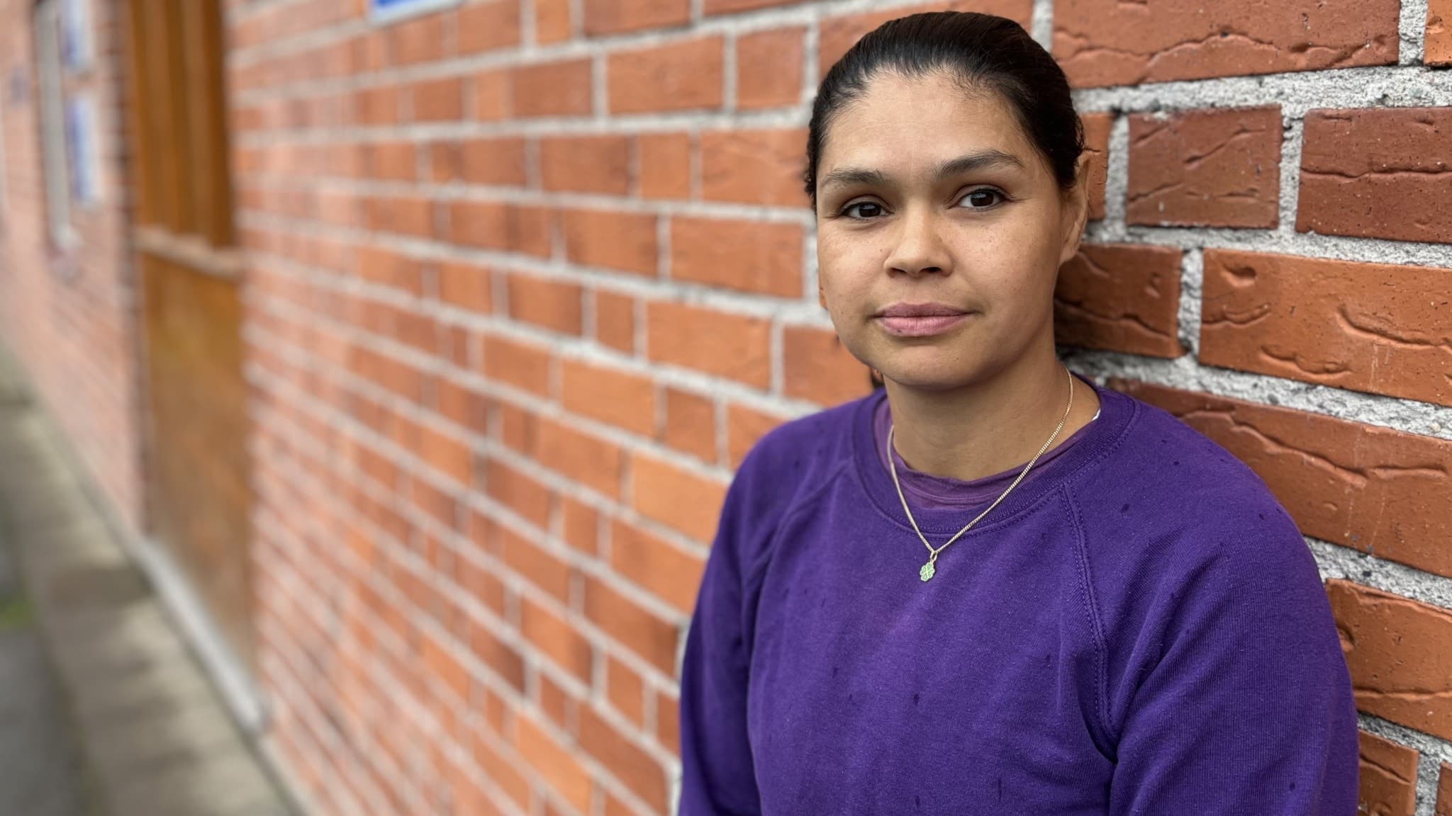 En kvinna i lila tröja står lutad mot en tegelvägg. Hon är lärare med 14 års erfarenhet och har nyligen utsatts för ett hot från en elev under sin graviditet.