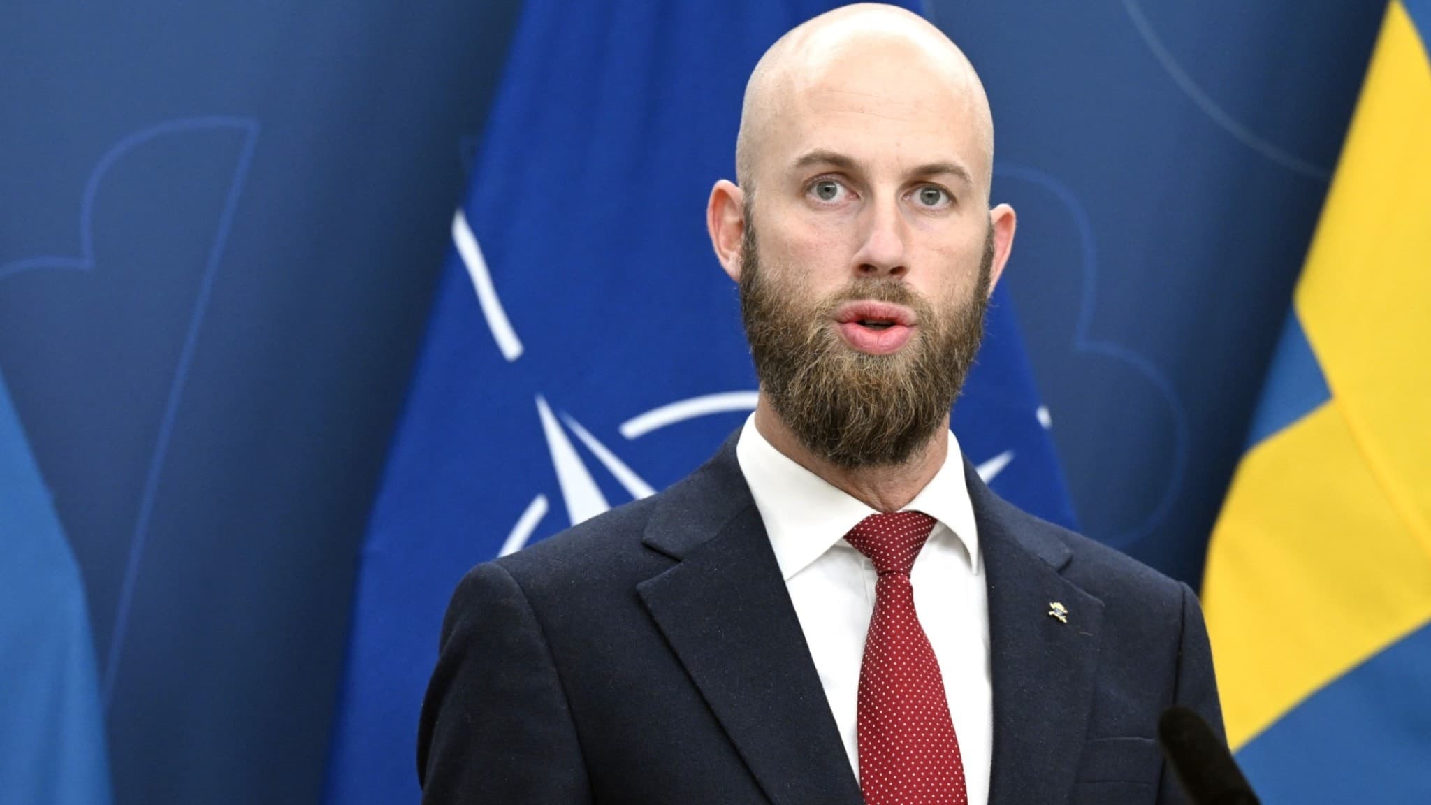 Carl-Oskar Bohlin (M), minister för civilt försvar, håller en presentation om satsningar på det civila försvaret. Han står vid ett podium framför svenska flaggor och en NATO-flagga.