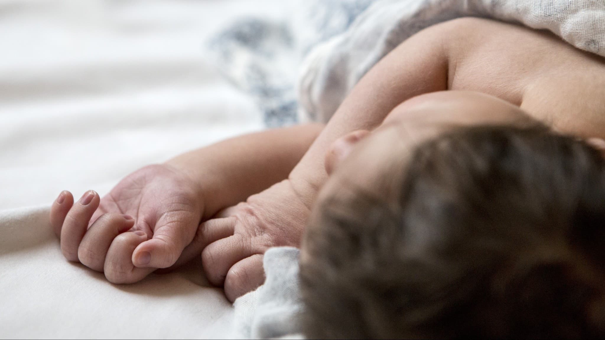 En nyfödd bebis ligger insvept i en filt på en ljus säng. Bebisen har mörkt hår och små händer som vilar avslappnat. Bilden symboliserar livets början och trygghet.