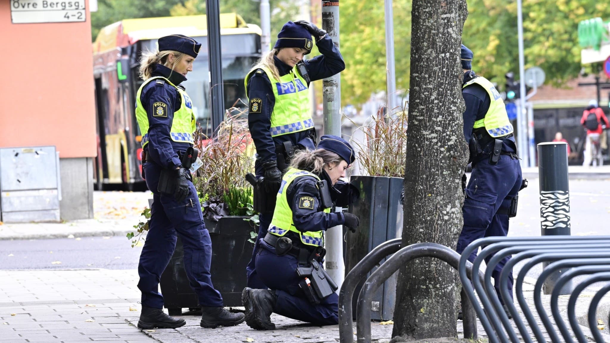 Fyra poliser undersöker en brottsplats i centrala Gävle efter en skottlossning där sex personer skadats. Området är omgivet av butiker, restauranger och stadsmiljö.