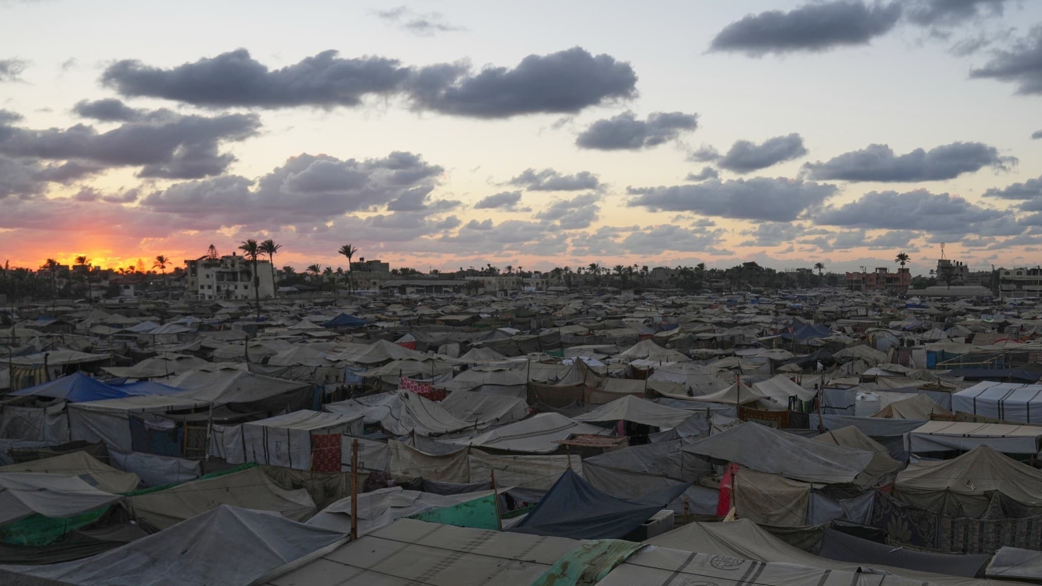 Solnedgång över ett stort tältläger för fördrivna palestinier i Deir al-Balah, centrala Gaza, med tält som breder ut sig under en himmel fylld av moln och palmträd i bakgrunden.