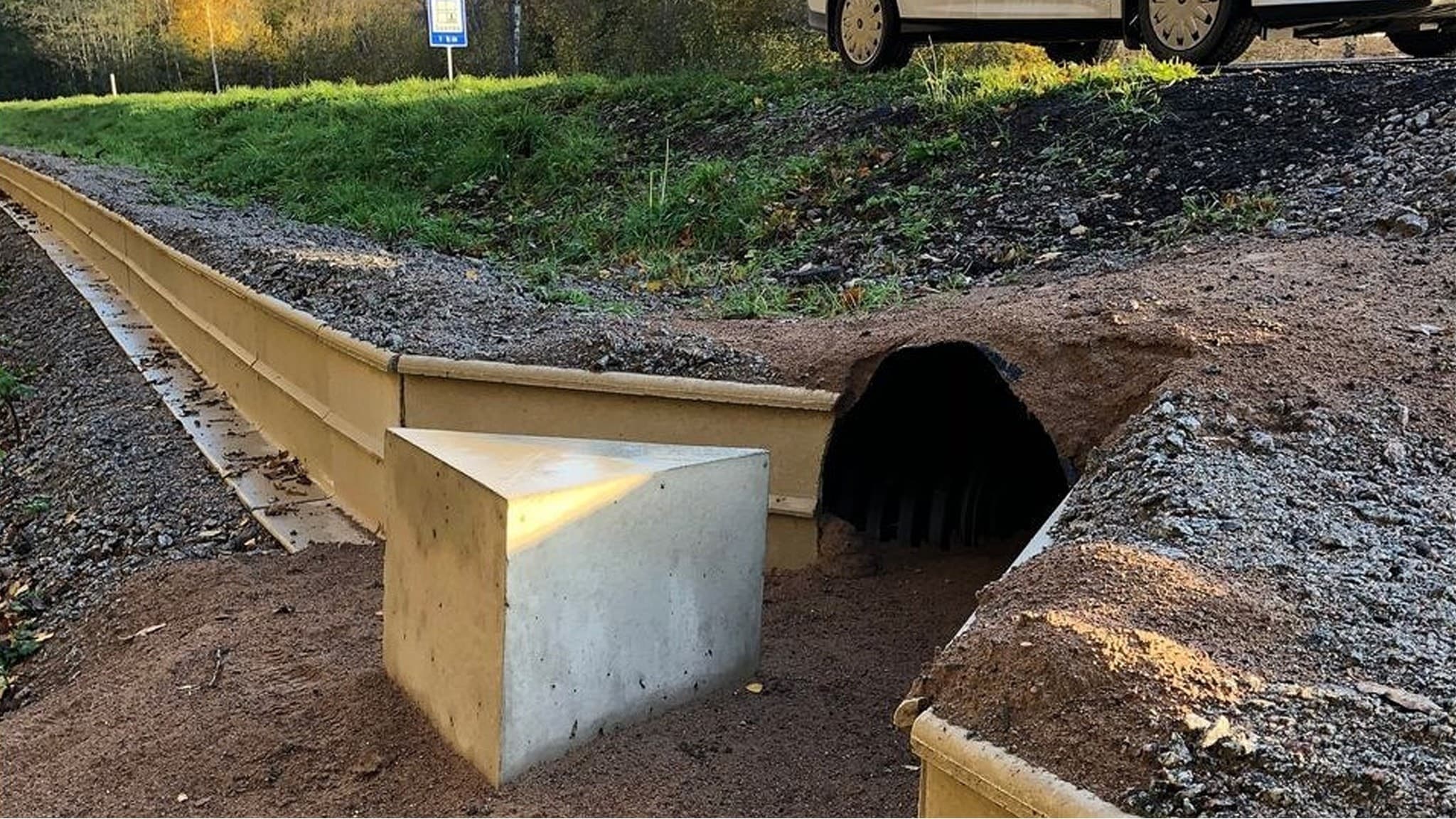En väldigt liten vägtrumma under en grusväg, omgiven av gräs och grus. Ett betongblock markerar trummans ingång, och en gul linje är synlig tvärs över området.