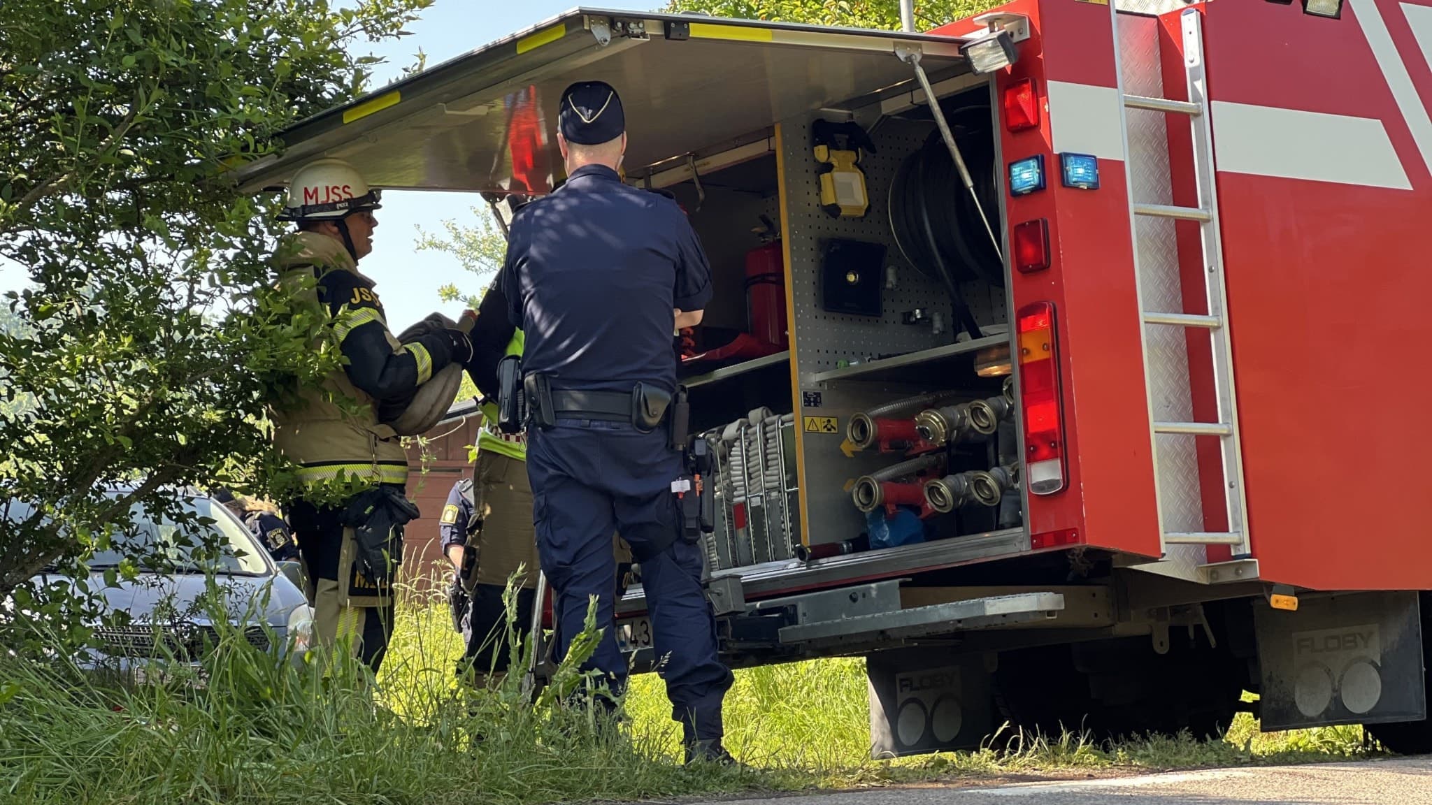 Brandmän och polis vid en brandbil, öppet lastutrymme med utrustning syns. Solig dag, gröna träd i bakgrunden. Räddningsinsats pågår.