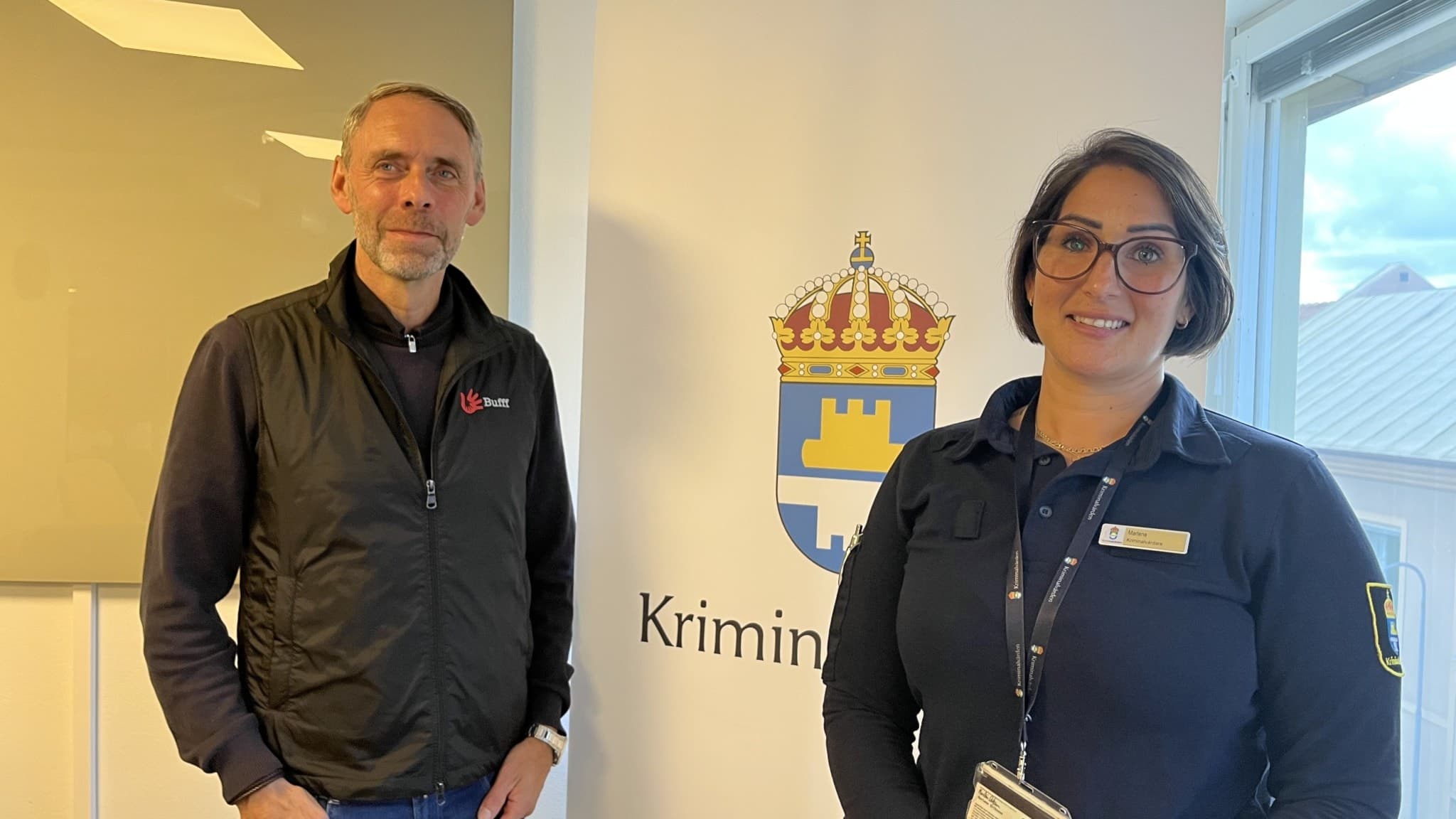 Två personer står framför en skylt med Kriminalvårdens logotyp. Bakgrunden visar ett kontorsfönster och en ljus interiör. Personerna bär arbetskläder med synliga namnbrickor.