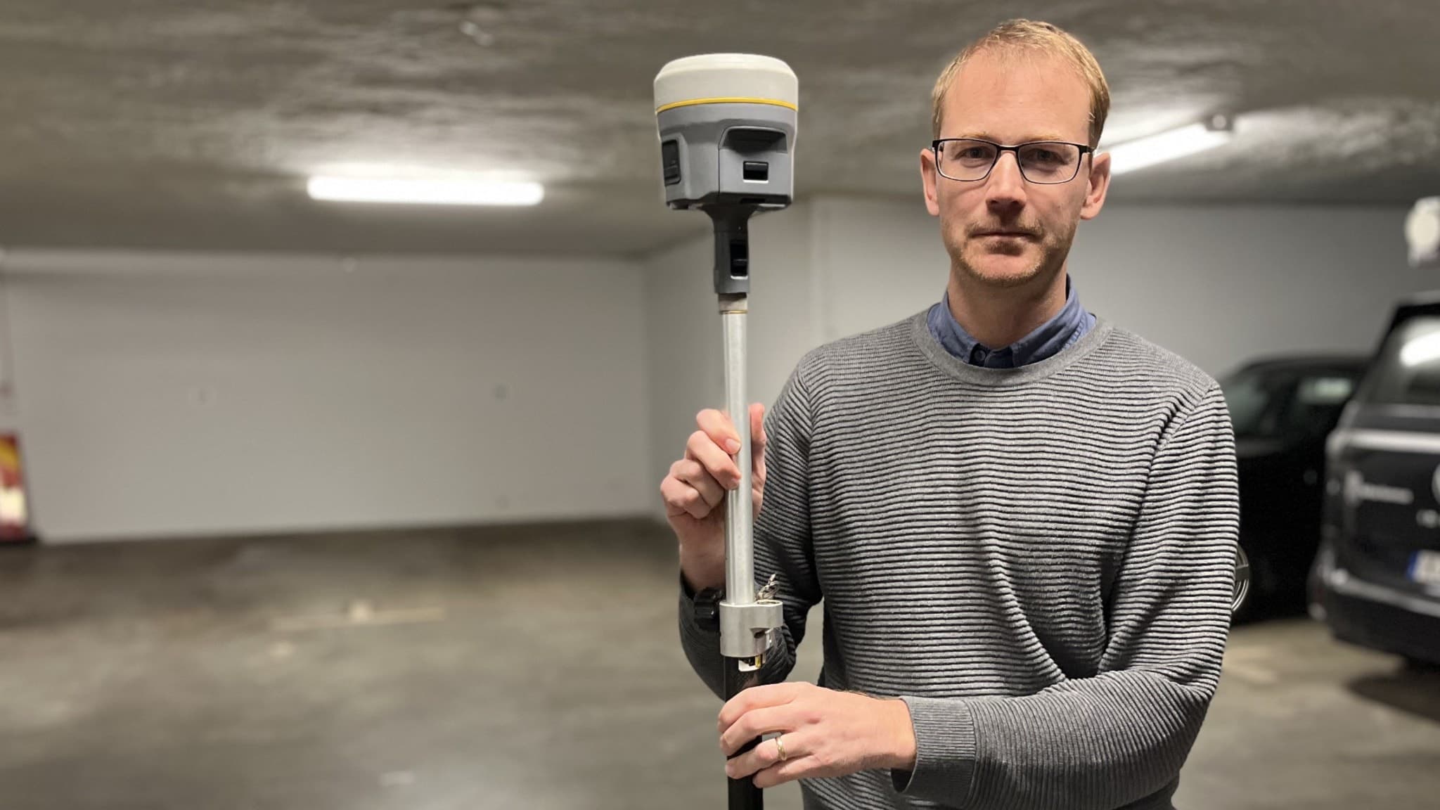 En person håller i en geodetisk mätutrustning i ett garage med parkerade bilar i bakgrunden. Utrustningen används för precisionsmätning inom kartläggning och byggprojekt.