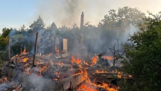 Ett hus har brunnit ner till grunden, omgivande vegetation syns i bakgrunden medan lågorna fortfarande brinner bland resterna. Röken stiger mot en klar himmel.