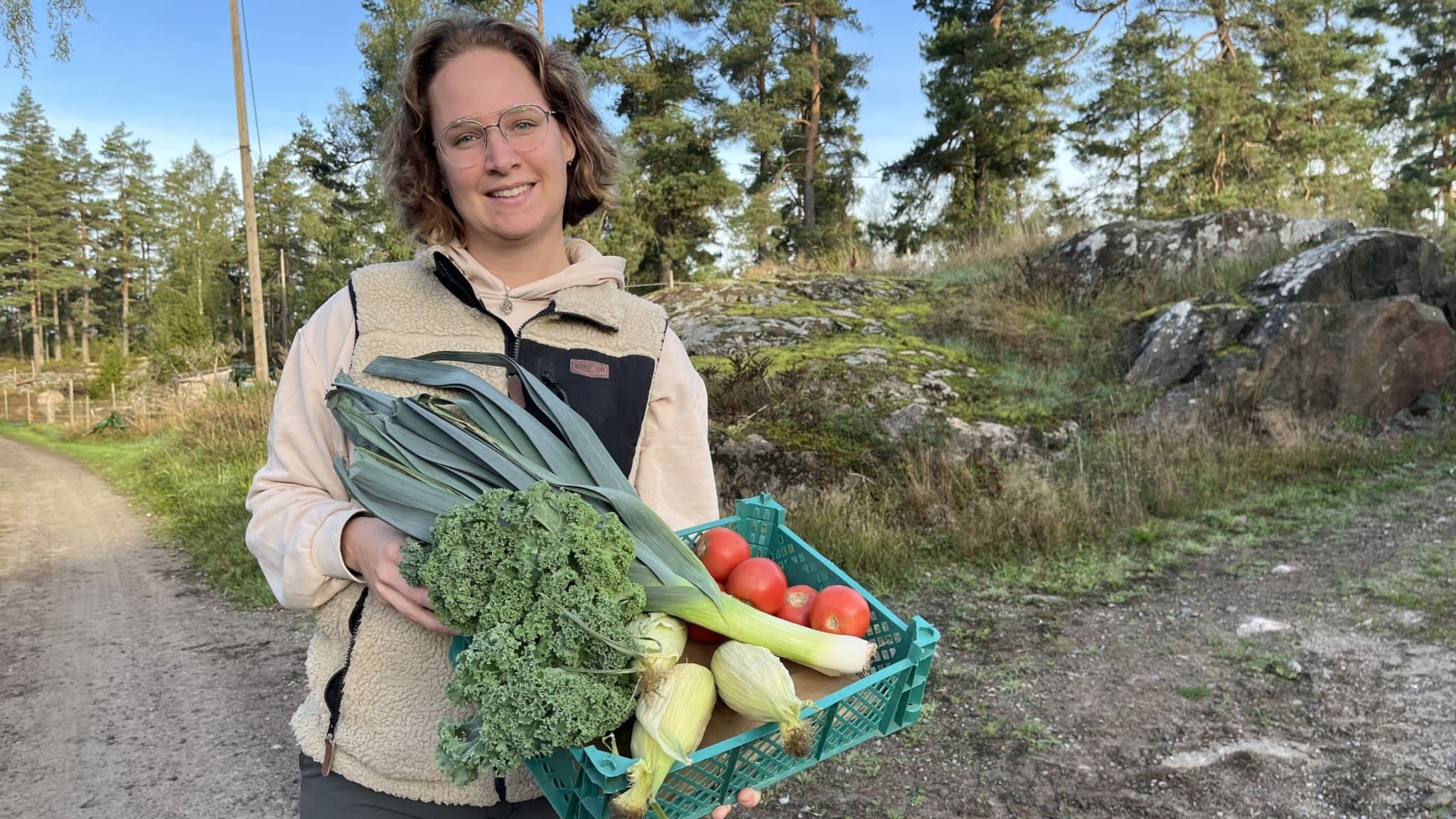 Ellen Olovsson står utomhus på en grusväg och håller en grön korg fylld med grönsaker som purjolök och tomater . Hon är omgiven av skog och lantlig miljö.
