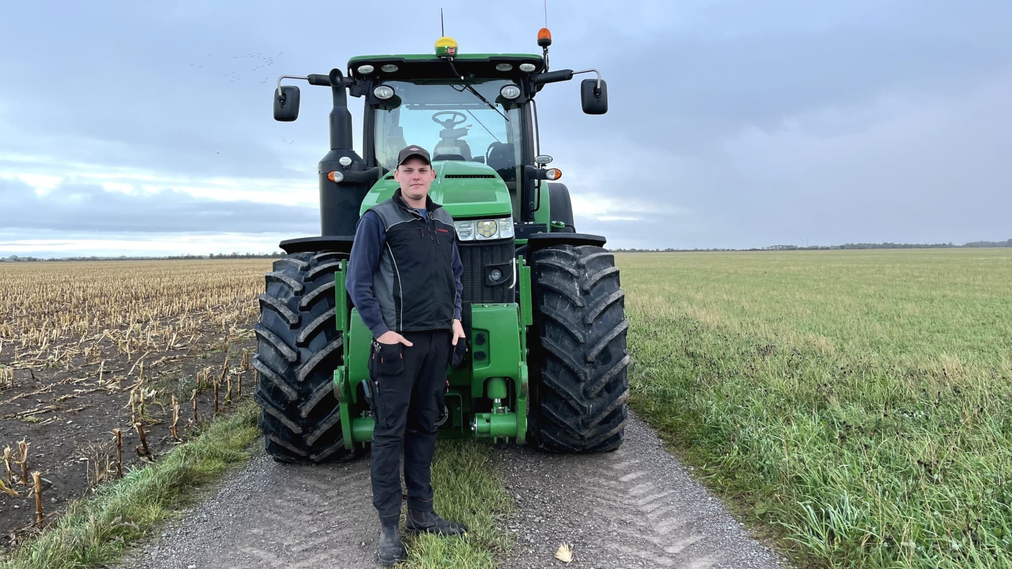 Axel Wiström står framför en grön traktor på en smal grusväg omgiven av åkrar under en mulen himmel. Jordbrukslandskapet sträcker sig långt i bakgrunden.