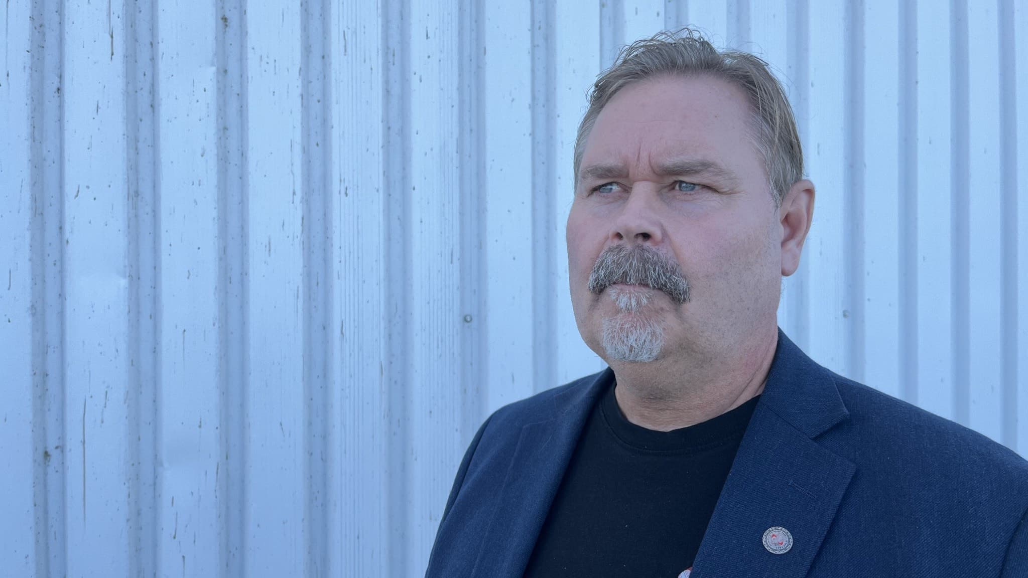 Håkan Bergenhav från IF Metall står framför en ljusgrå korrugerad plåtvägg, klädd i en blå kavaj med en nål på slaget.