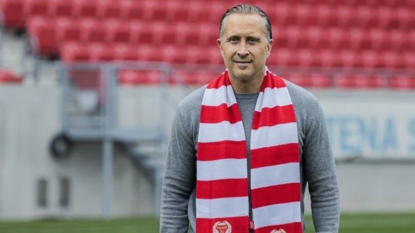En man står på fotbollsplanen med en rödvit halsduk runt halsen. Bakgrunden visar läktare med röda stolar.