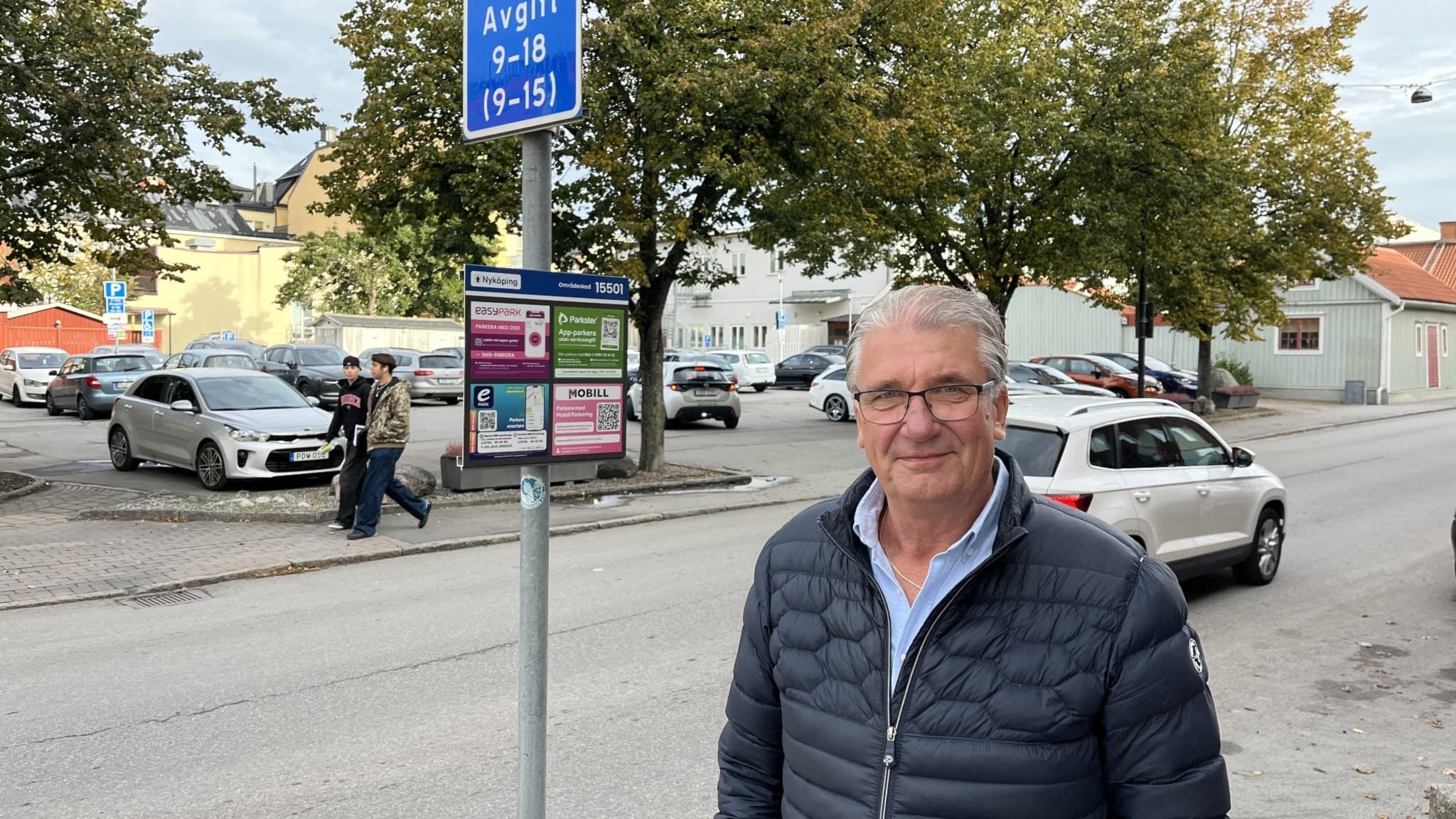 Anders Isaksson står vid en parkeringsskylt på en trafikerad gata med träd i bakgrunden. Flera bilar kör förbi medan några personer går längs trottoaren.