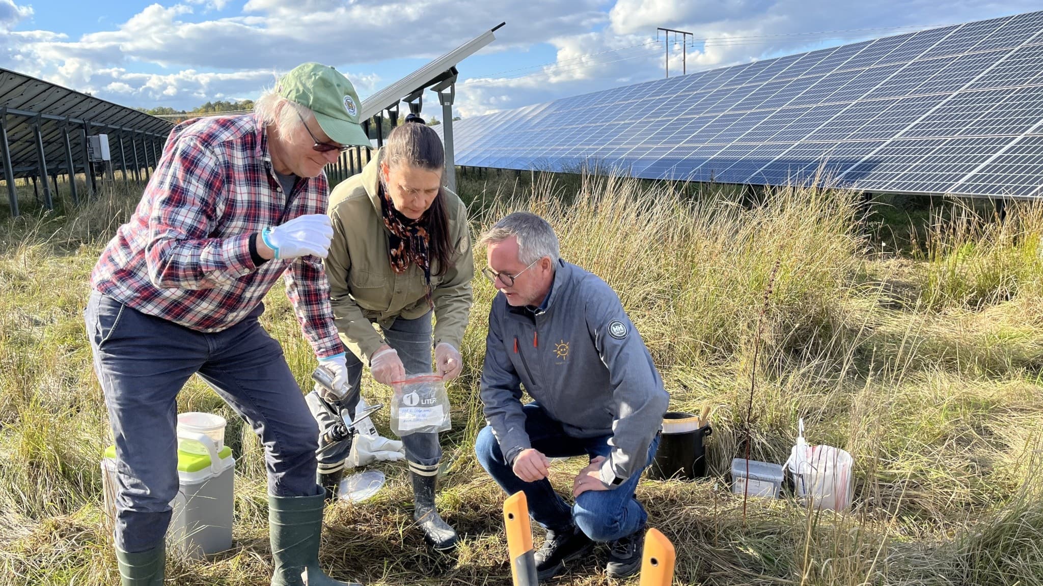 Tre forskare undersöker markprover i en solcellspark omgivna av stora solpaneler under en klar himmel, vilket indikerar ett forskningsprojekt inom förnybar energi.