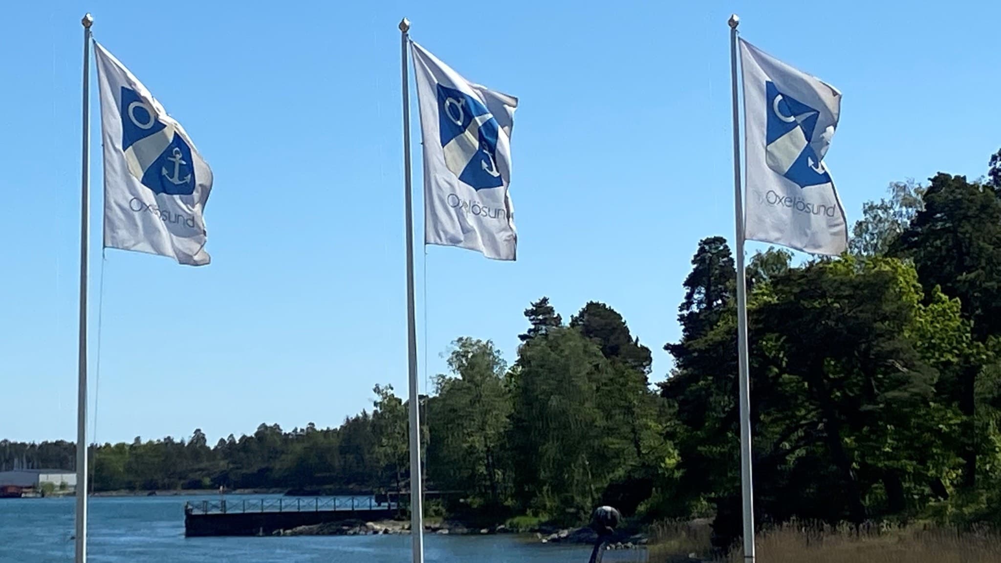 Tre flaggor med blå emblem står vid ett vatten, omgivna av grönskande träd och en brygga i bakgrunden under en klarblå himmel.