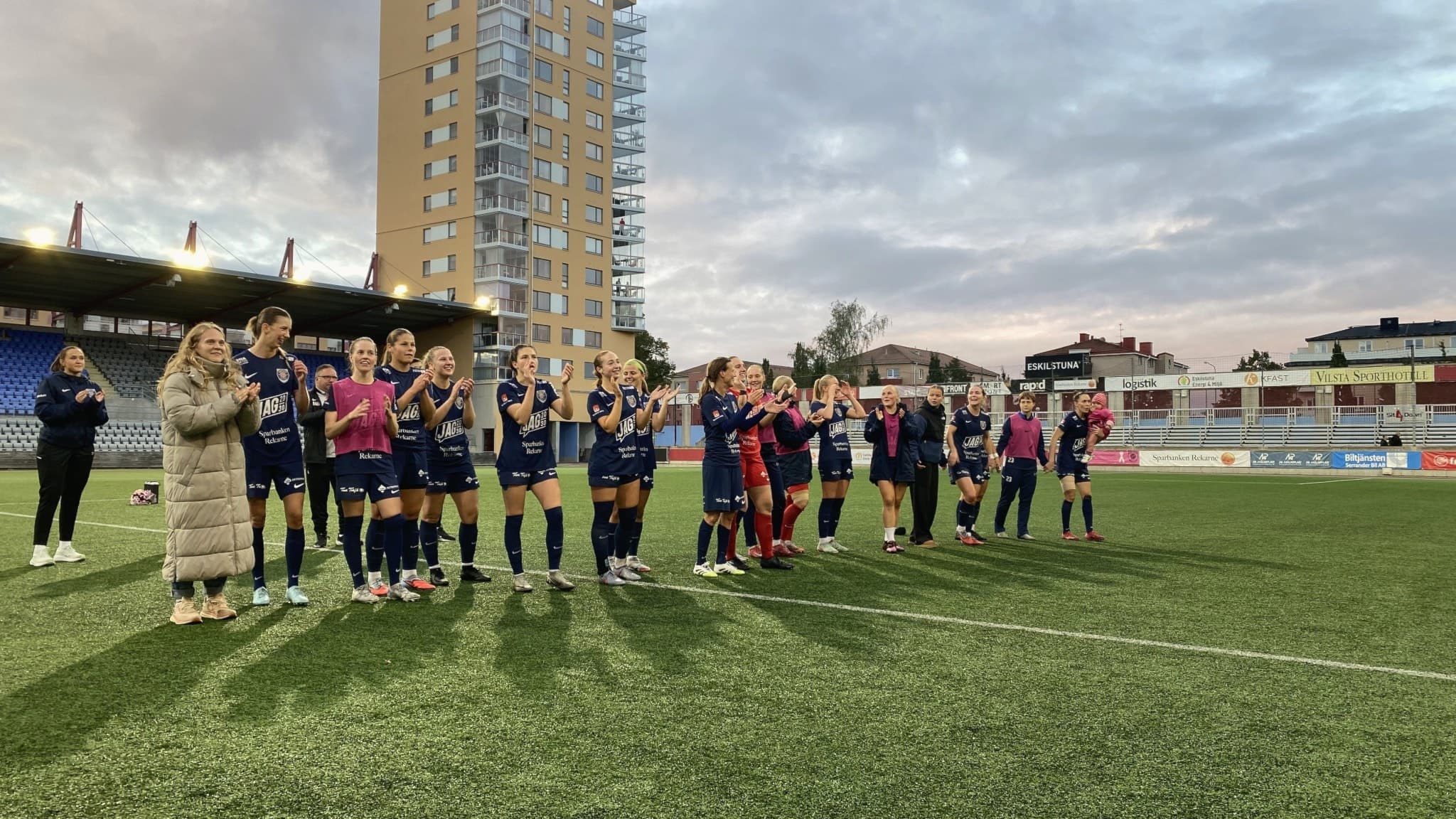 Fotbollsspelare från Eskilstuna United står på rad på en konstgräsplan, omgivna av höga byggnader och läktare, med kvällshimlen i bakgrunden. Laget verkar delta i en ceremoni eller hyllning.